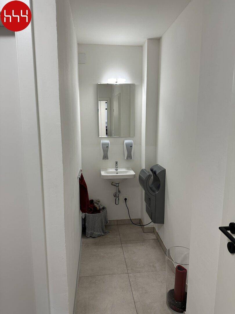 Schmaler, moderner Toilettenraum mit Waschbecken, Spiegel, Seifenspendern und einem schnellen Händetrockner.