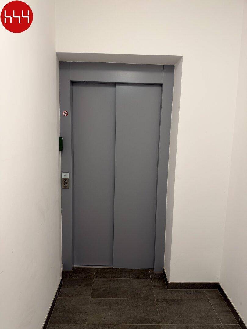 Moderner Aufzug im Flur eines Wohngebäudes mit grauen Schiebetüren und einfacher Ausstattung.