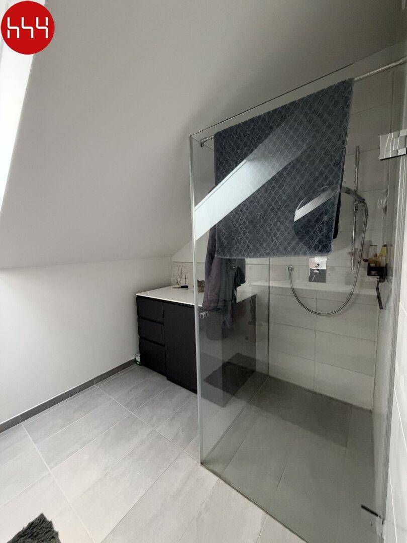 Modernes Badezimmer mit begehbarer Dusche, dunklem Waschtisch und hellen Fliesen unter der Dachschräge.