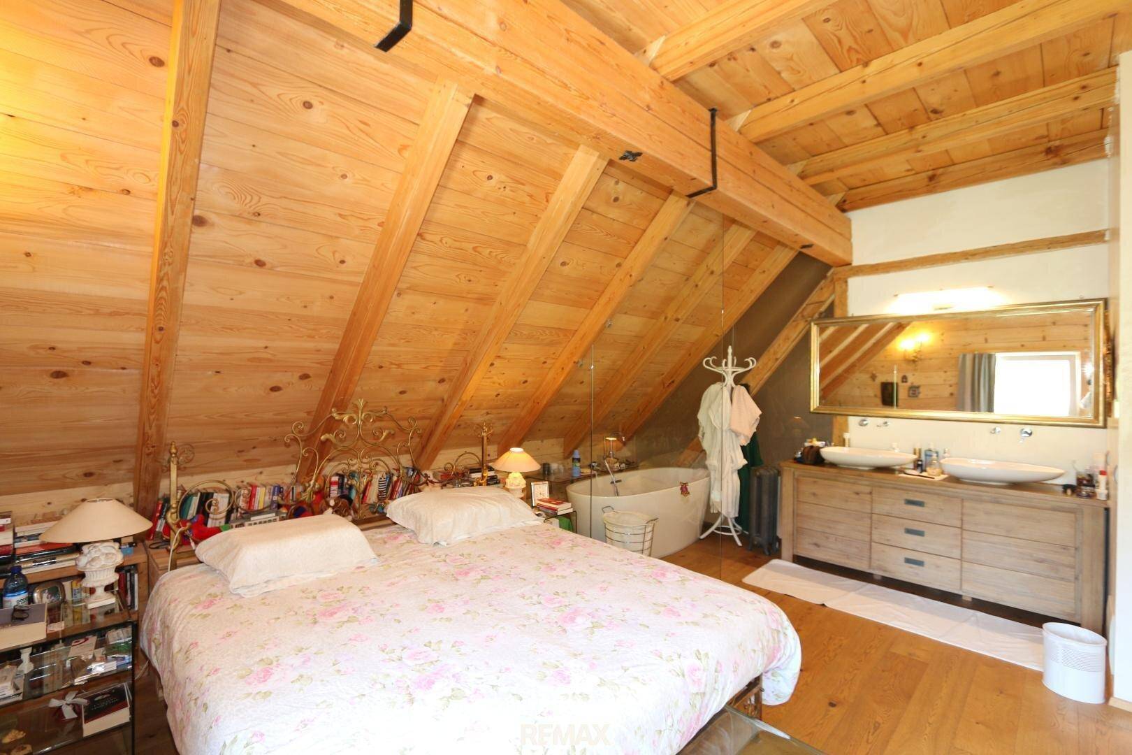 Großes Schlafzimmer mit integriertem Badebereich, freistehender Badewanne und Doppelwaschbecken unter Holzbalken.