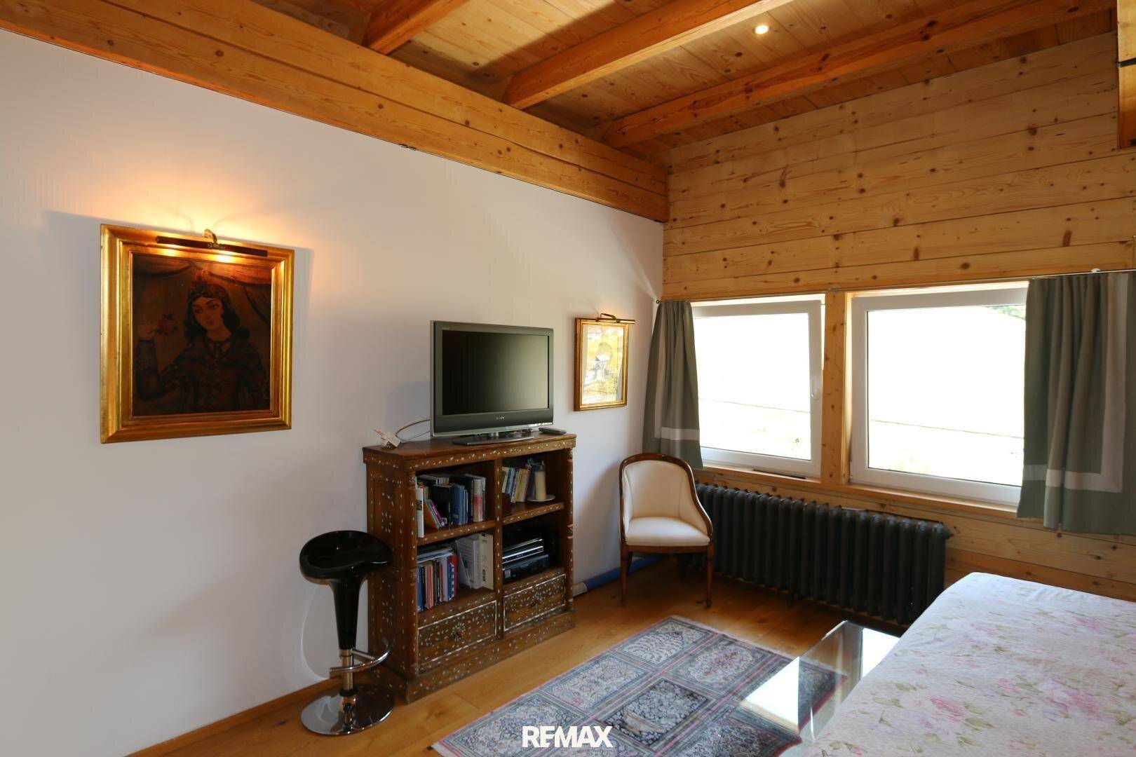 Helles Zimmer mit Holzwänden, freiliegenden Balken und einem Fenster, ausgestattet mit TV und Bücherregal.