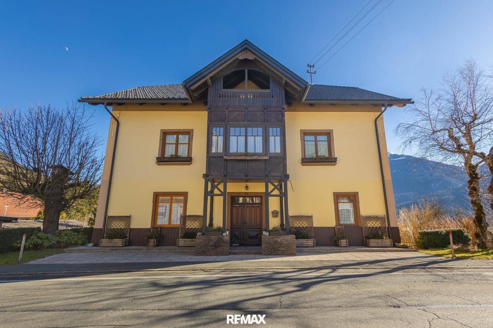Traditionelles Haus mit gelber Fassade, Holzbalkon und Bergblick unter blauem Himmel.