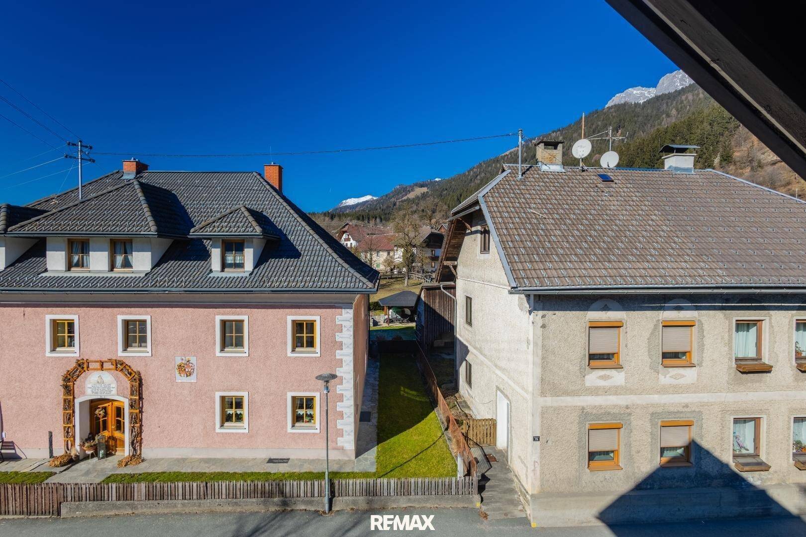Zwei traditionelle Häuser in ländlicher Umgebung mit Bergen im Hintergrund unter blauem Himmel.