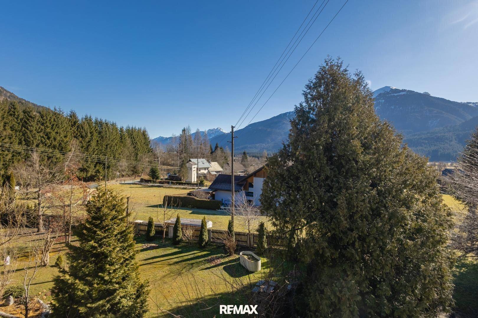 Weitläufige grüne Landschaft mit Bergen im Hintergrund und vereinzelten Häusern unter blauem Himmel.