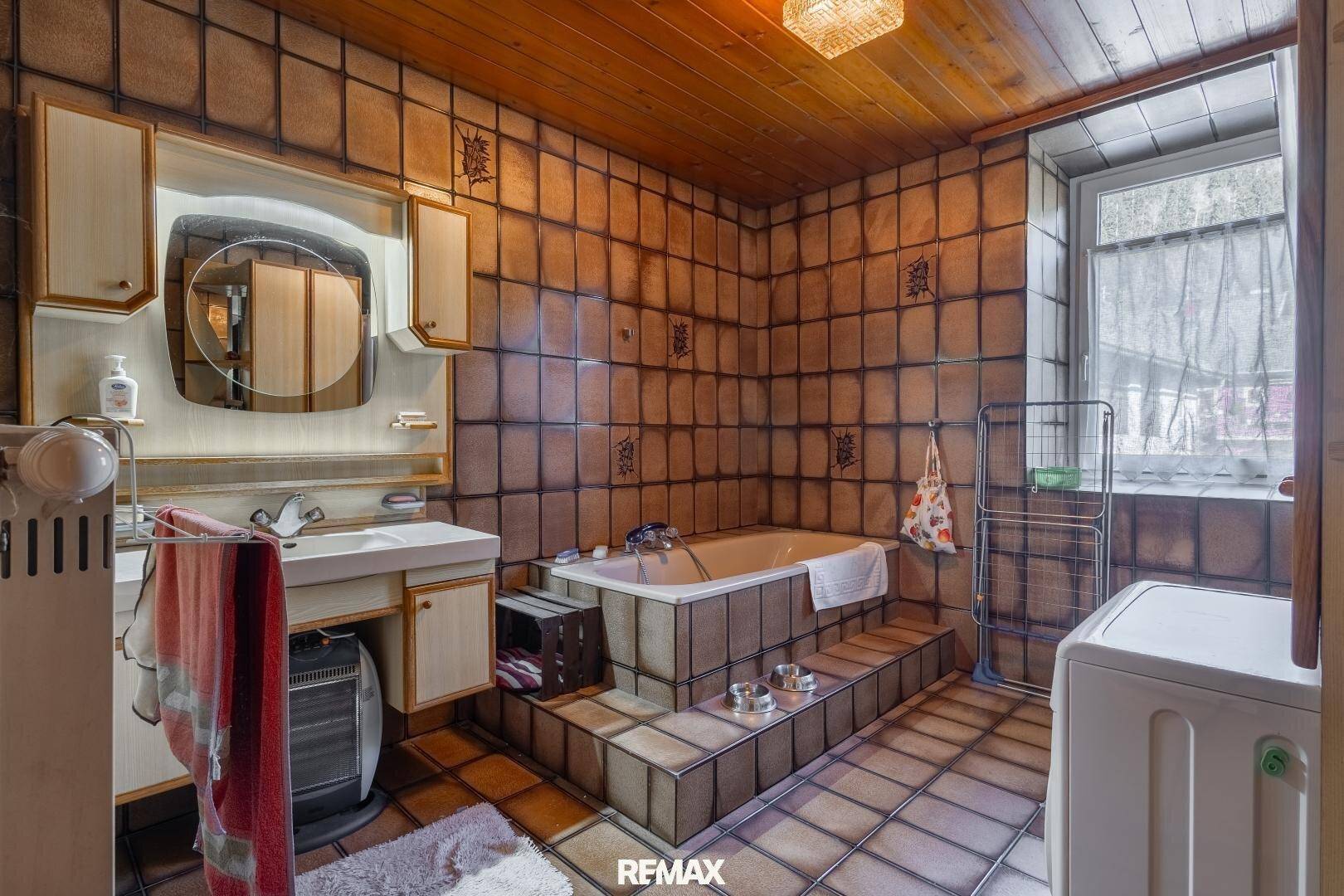 Rustikales Badezimmer mit braunen Fliesen, Holzdecke und Badewanne, ausgestattet mit Waschmaschine.