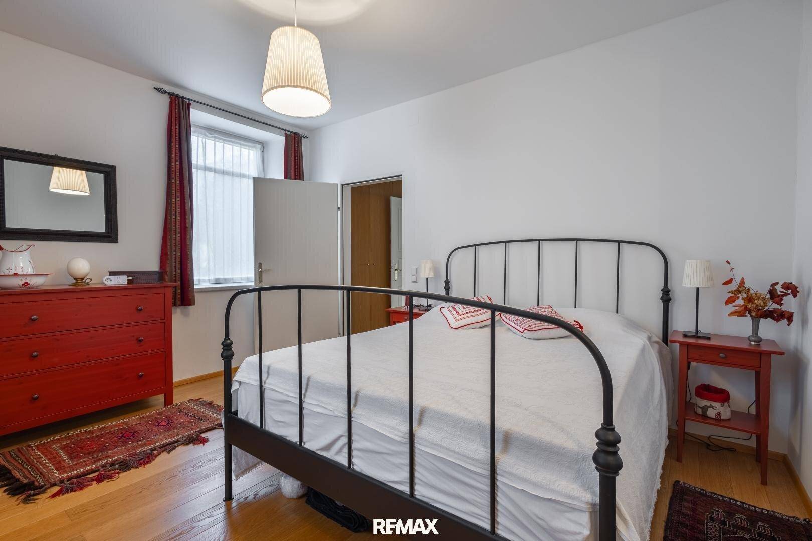 Helles Schlafzimmer mit Holzboden, schmiedeeisernem Bett und roten Akzenten, gemütlich eingerichtet.