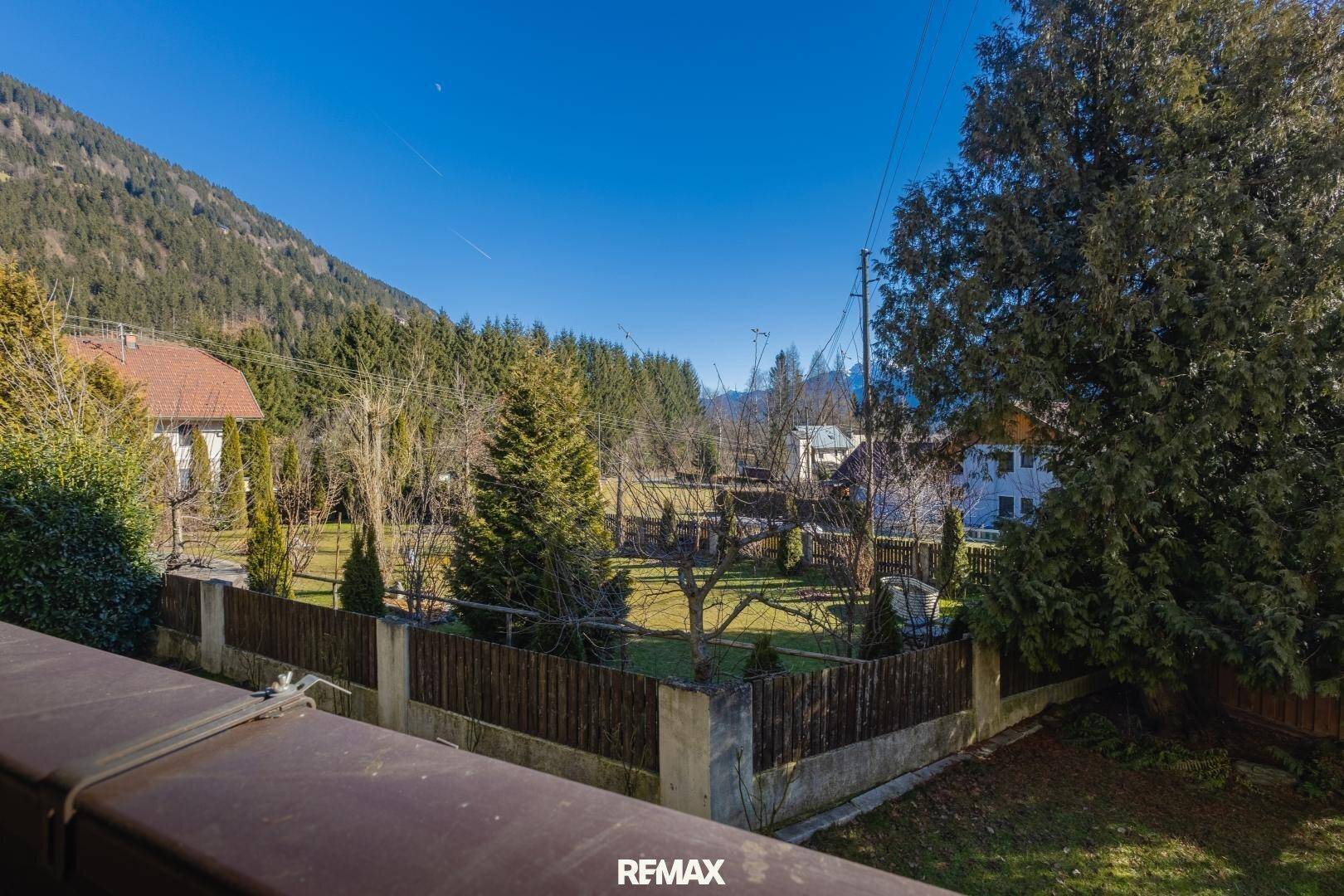 Blick vom Balkon auf einen gepflegten Garten mit Bäumen und die umliegende Berglandschaft.