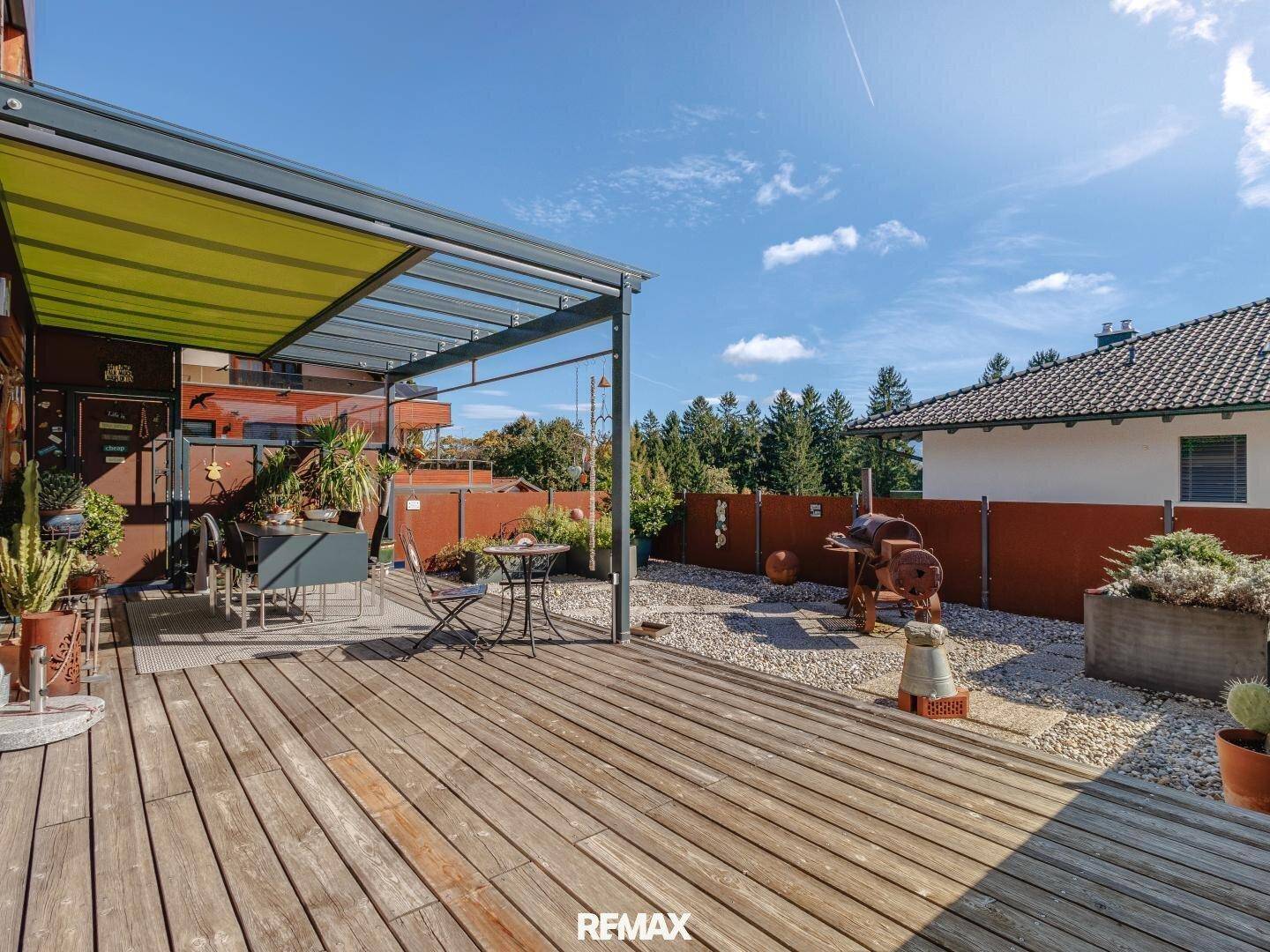 Große Holzterrasse mit Überdachung und gemütlichem Sitzbereich, umgeben von Kies und Pflanzen.
