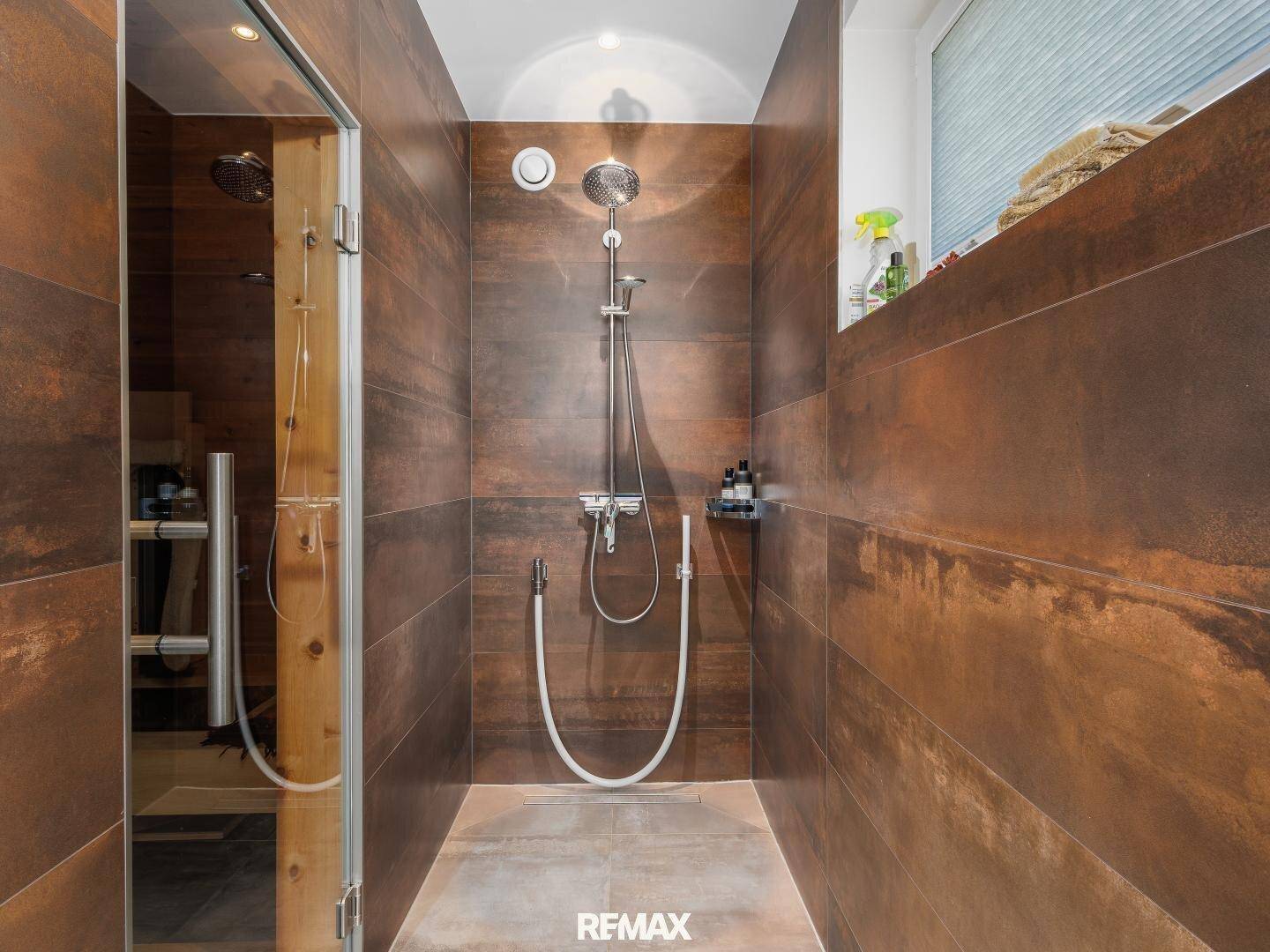 Geräumige Dusche mit braunen Fliesen und direktem Zugang zu einer sichtbaren Sauna.