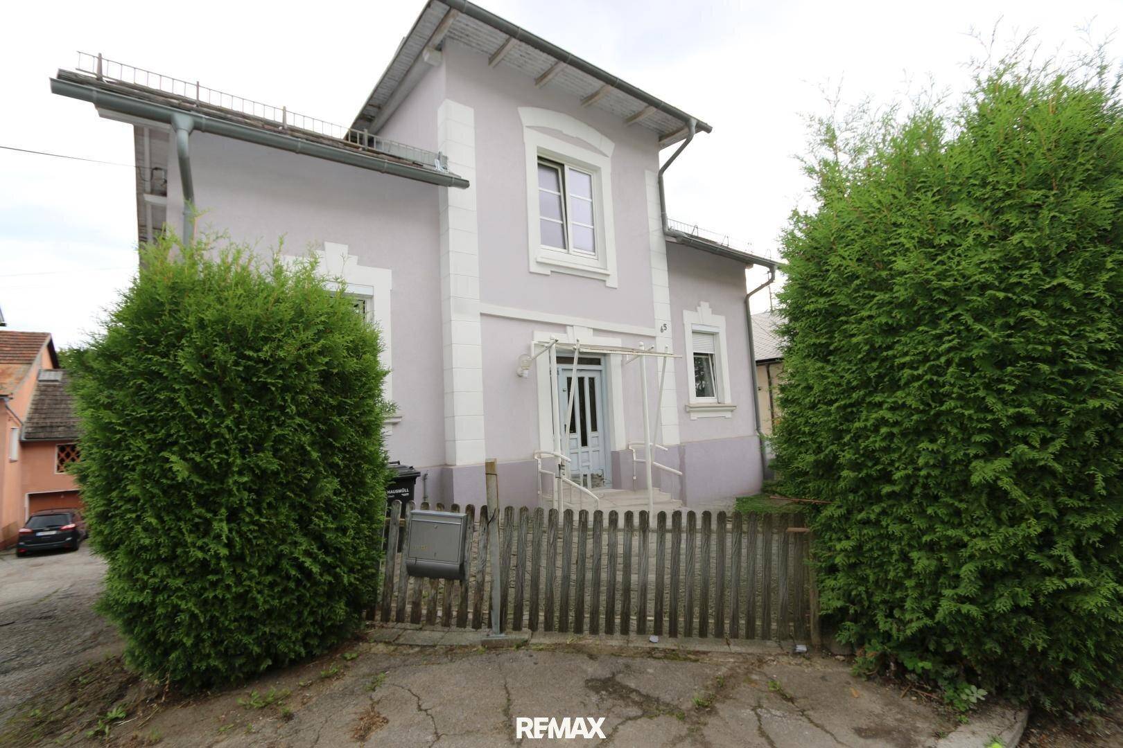 Ansicht eines hellgrauen Hauses mit weißer Fassade, umgeben von hohen Hecken und einem Holzzaun.