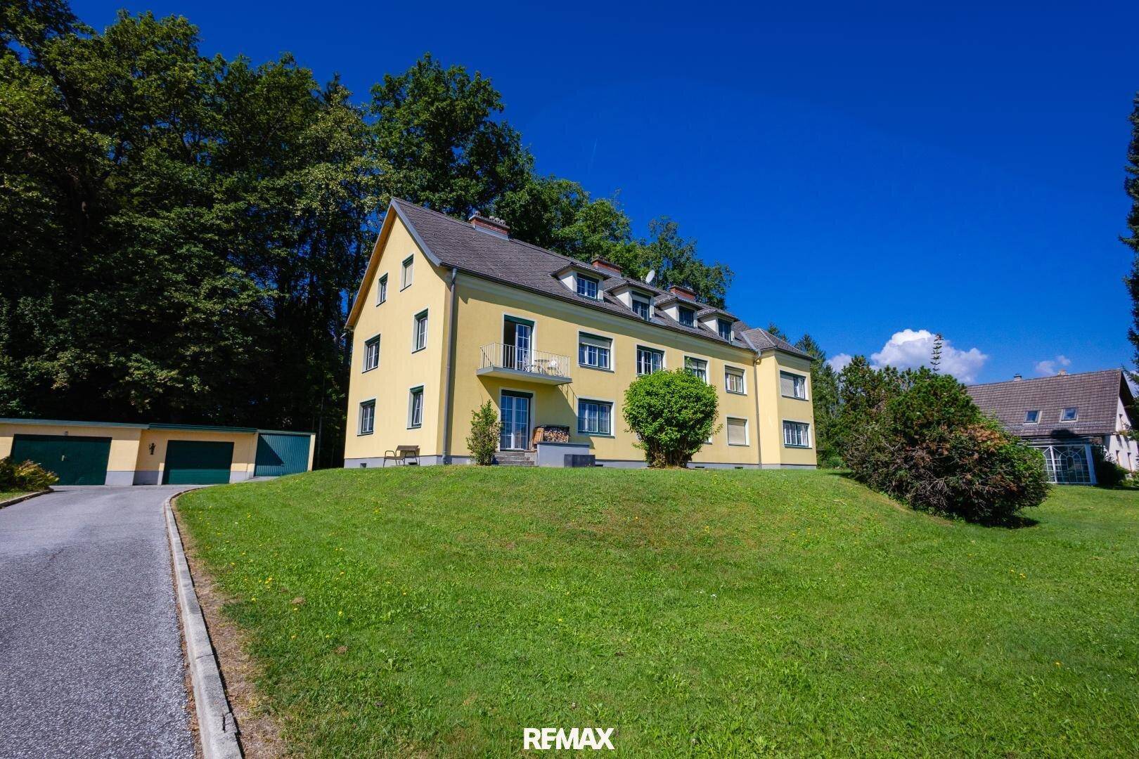 Gelbes Mehrfamilienhaus mit grüner Umgebung und Garagen im Hintergrund.