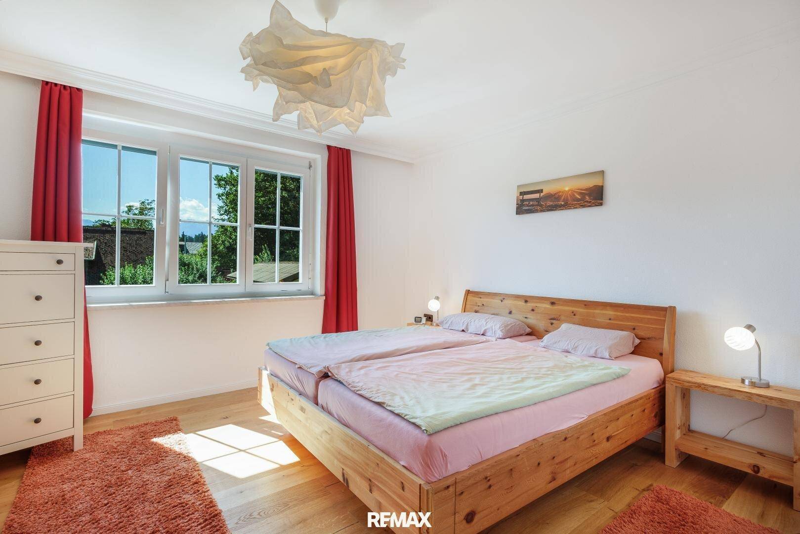 Helles Schlafzimmer mit Holzbett, weißer Kommode und roten Vorhängen, sonniger Ausblick ins Grüne.