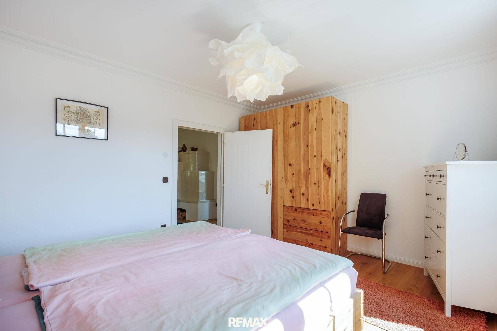 Schlafzimmer mit Holzbett und großem Holzschrank, Zugang zu einem weiteren Raum mit Kachelofen.