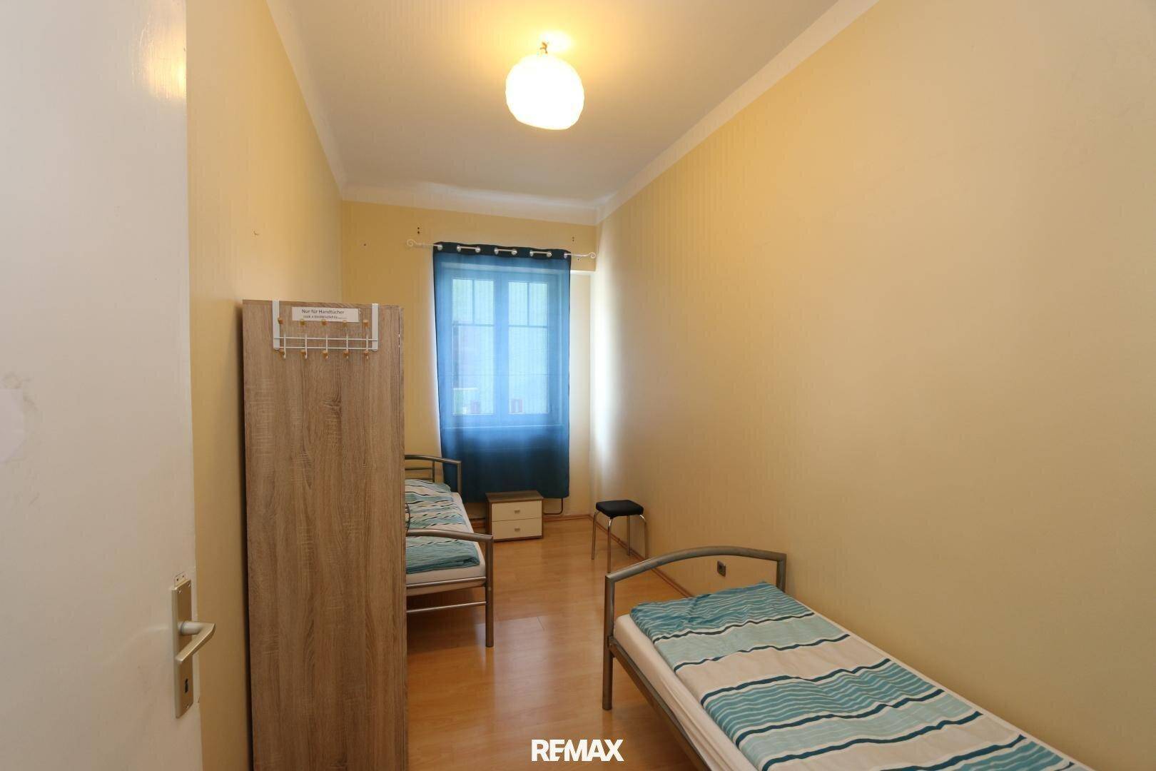 Schlafzimmer mit zwei Einzelbetten, hellgelben Wänden und einem Fenster mit blauen Vorhängen.