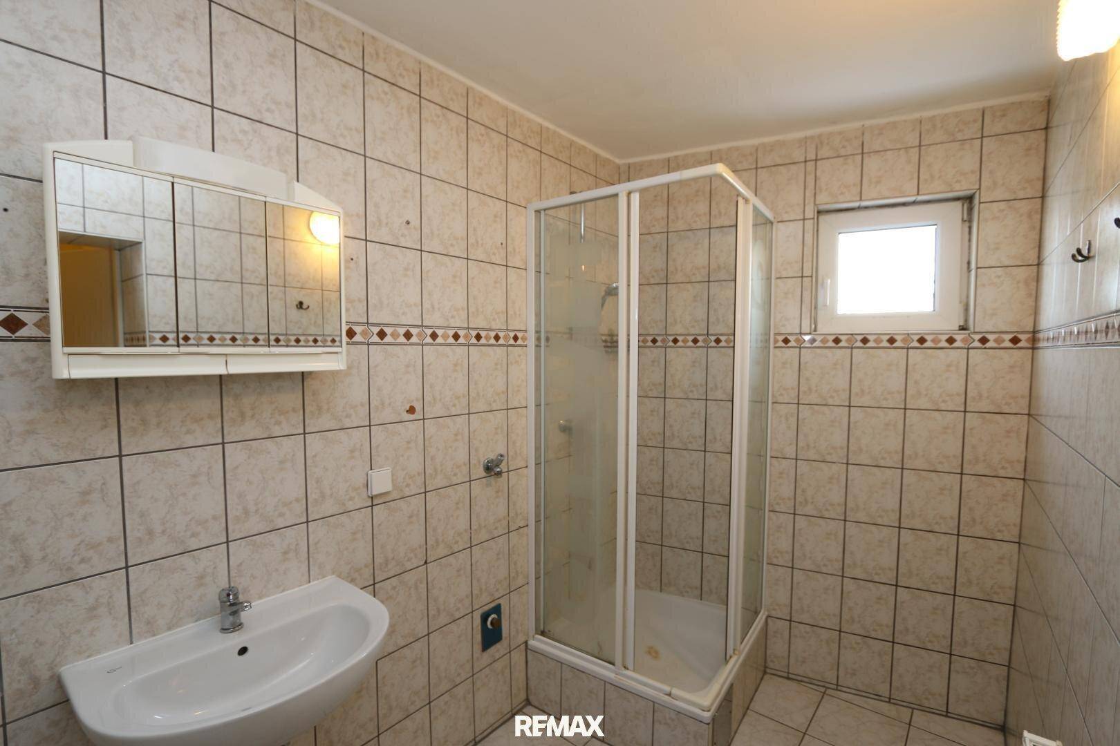 Badezimmer mit beigefarbenen Fliesen, einer Duschkabine, Waschbecken und einem Fenster für Tageslicht.
