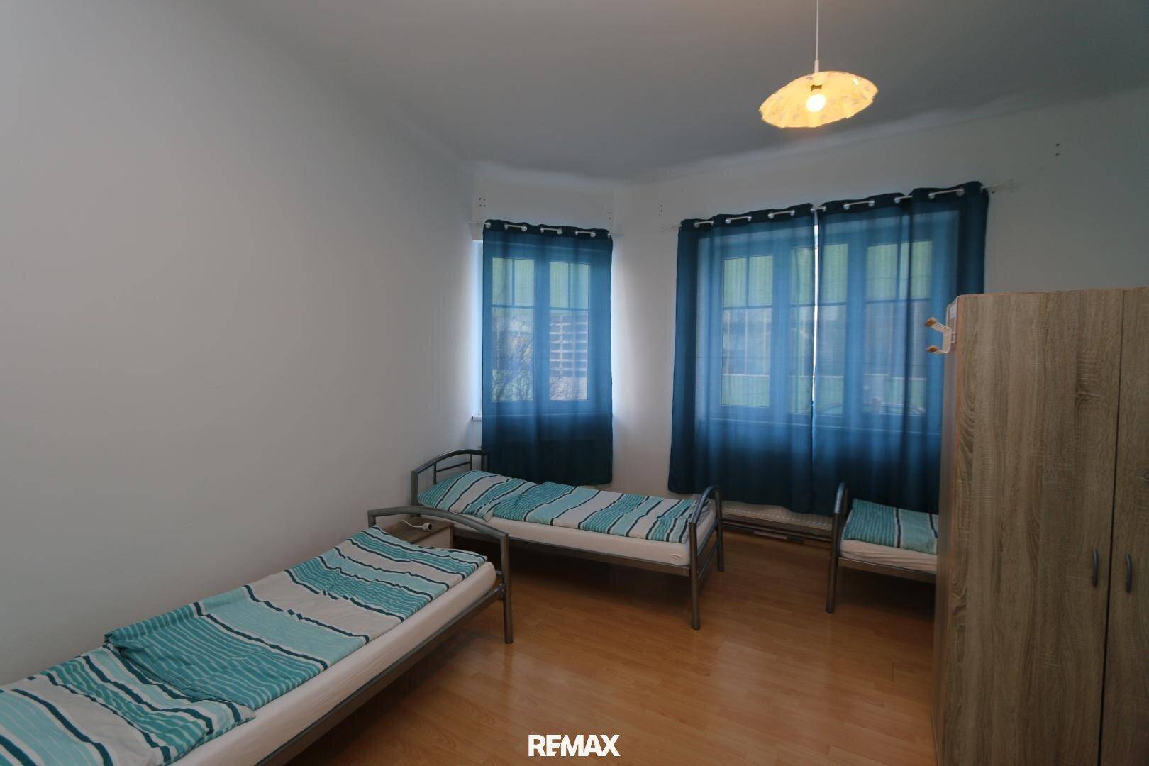 Schlafzimmer mit zwei Einzelbetten, weißen Wänden und zwei Fenstern mit blauen Vorhängen.