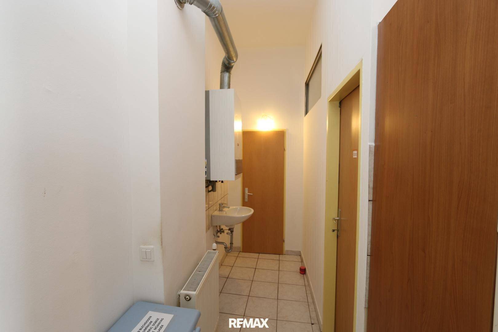 Schmaler Flur oder Hauswirtschaftsraum mit Waschbecken, Gastherme und Heizkörper, gefliestem Boden.