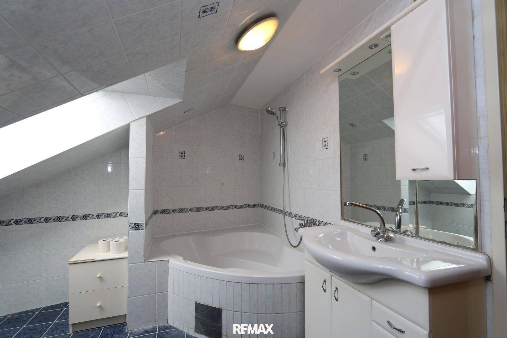 Dachgeschoss-Badezimmer mit Eckbadewanne, Waschbecken und schrägen Wänden, gefliest und mit Dachfenster.