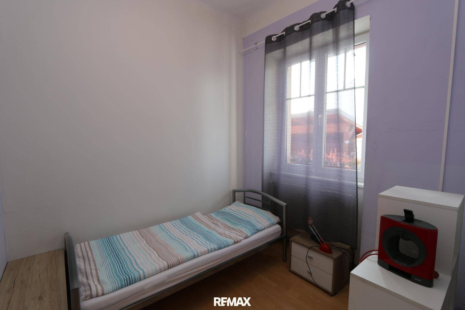 Schlafzimmer mit einem Einzelbett, lila und weißen Wänden und einem Fenster mit dunklen Vorhängen.