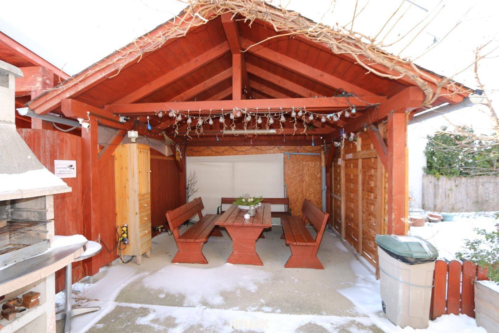 Überdachter Sitzbereich im Freien mit Holzbänken, Tisch und einem Grill, umgeben von Schnee.