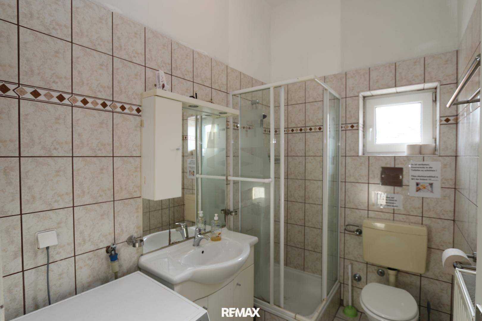 Gefliestes Badezimmer mit Duschkabine, WC, Waschbecken und Waschmaschine, inklusive Fenster für Tageslicht.