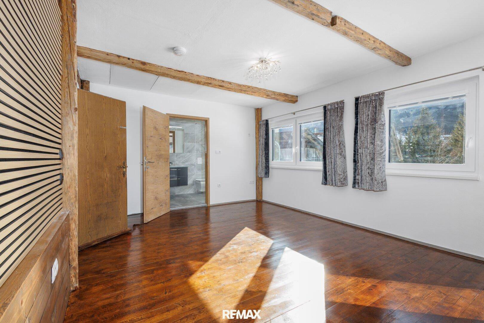 Schlafzimmer mit sichtbaren Holzbalken, Holzboden und Zugang zu einem angrenzenden Badezimmer.