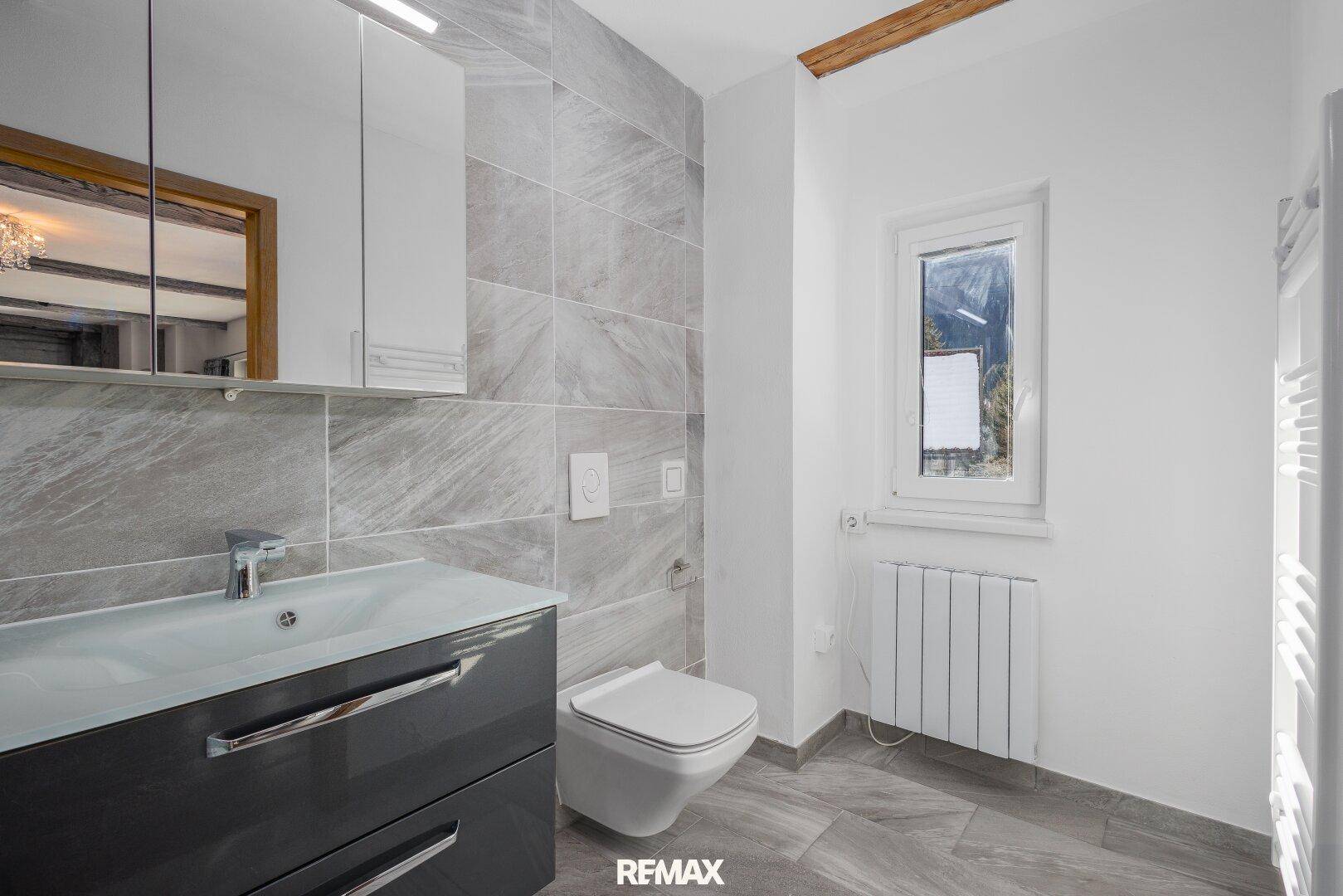 Helles Badezimmer mit modernen grauen Fliesen, WC, Waschtisch und Fenster für Tageslicht.