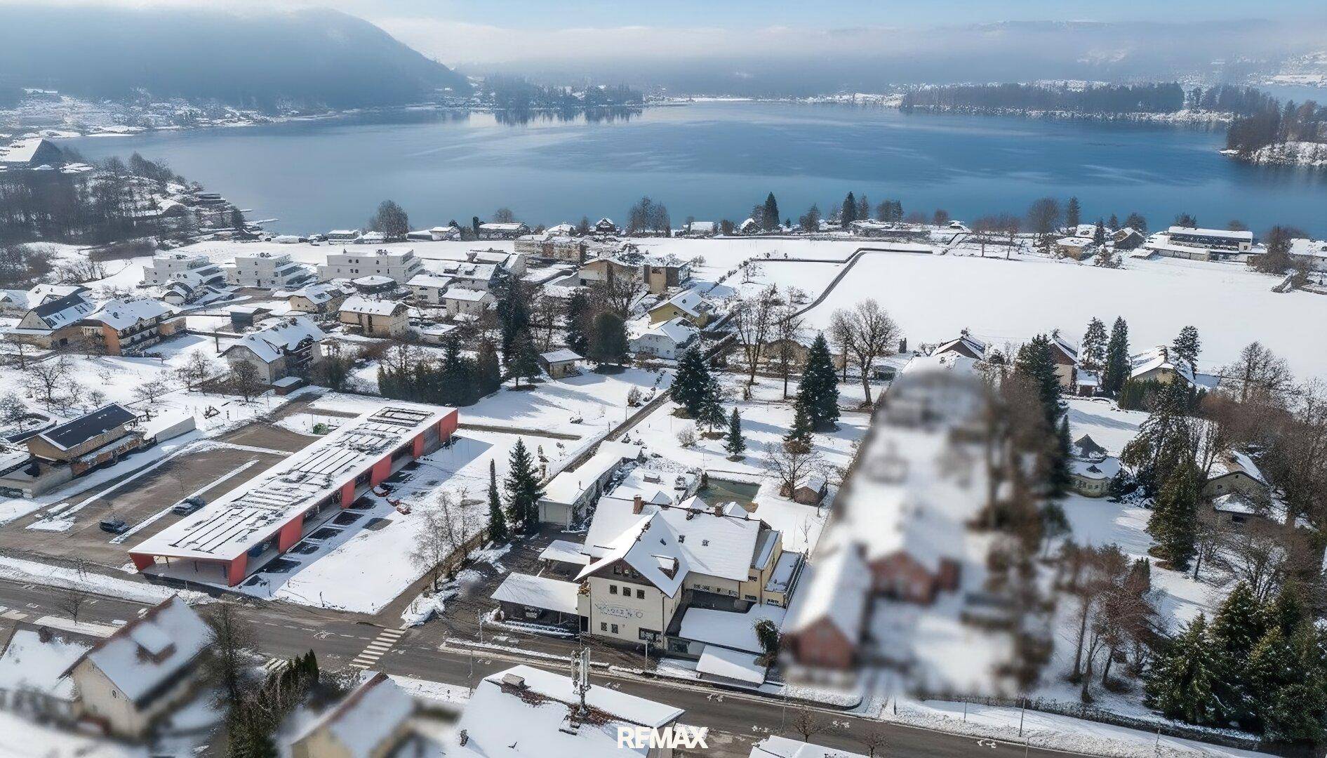 Winterliche Luftaufnahme einer Wohnsiedlung am Seeufer mit verschneiten Dächern und Bäumen.
