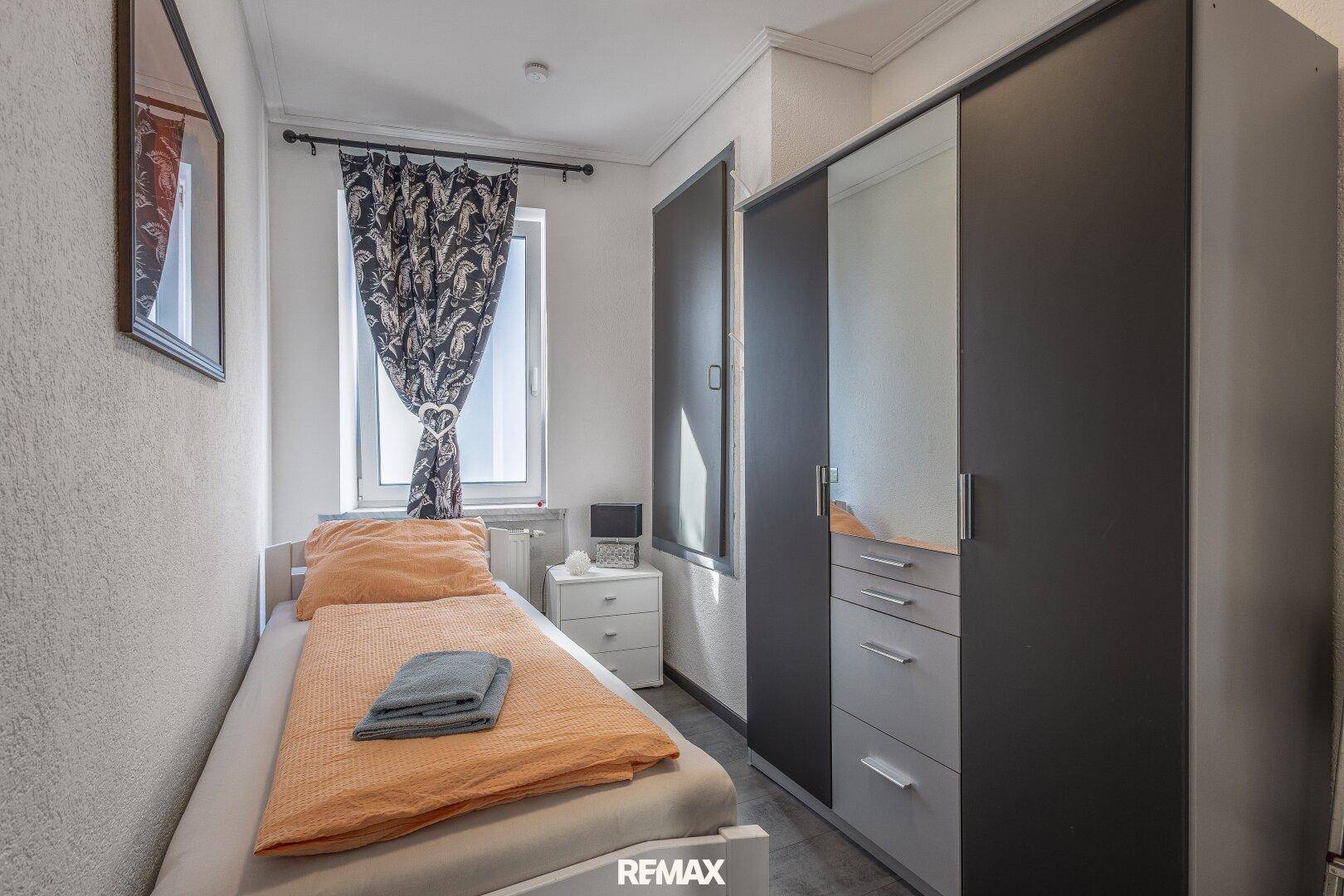 Kleines Schlafzimmer mit Einzelbett, Schrank und Fenster mit gemusterten Vorhängen.