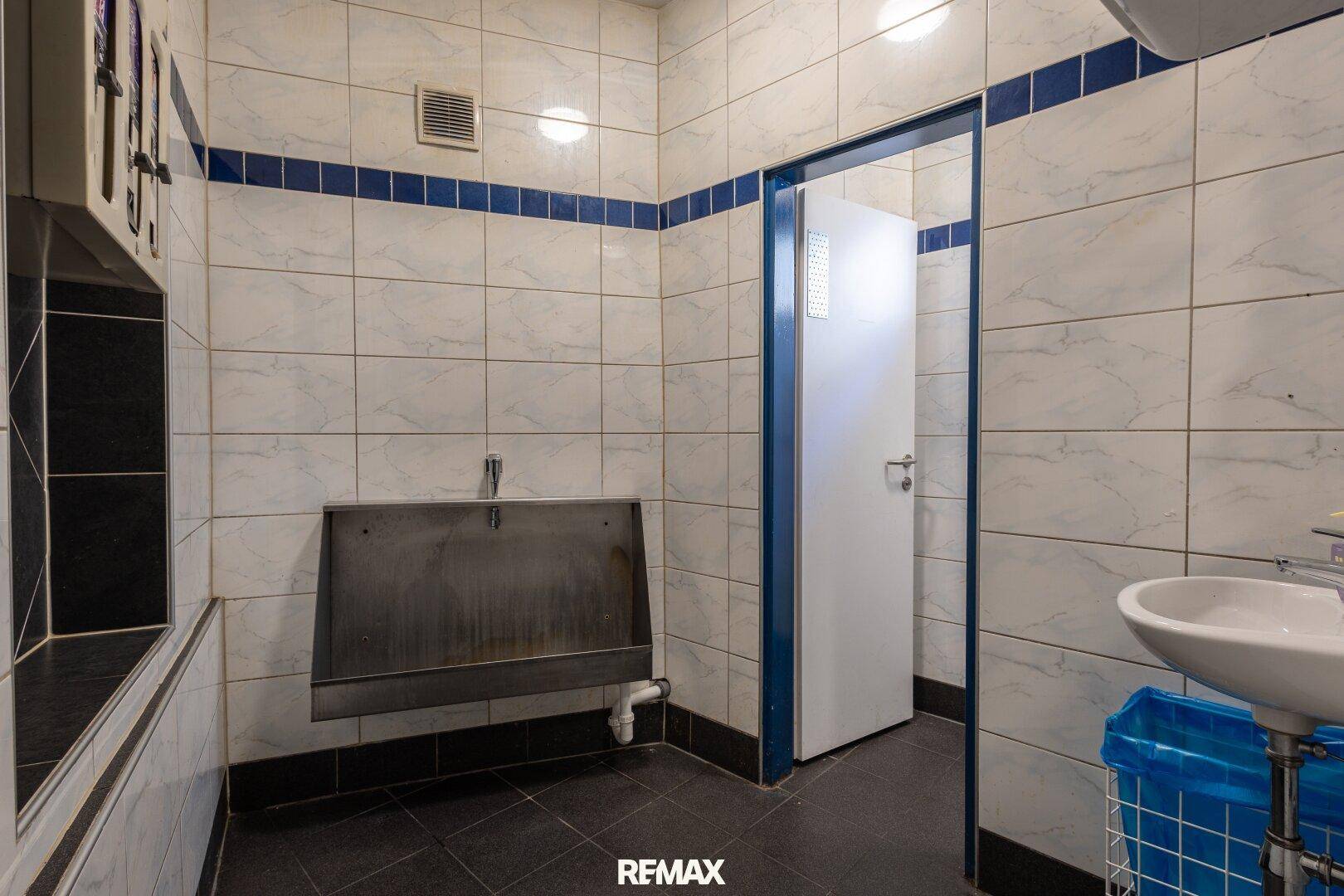 Gefliester Toilettenraum mit Urinal, Waschbecken und blauen Akzentstreifen an den Wänden.