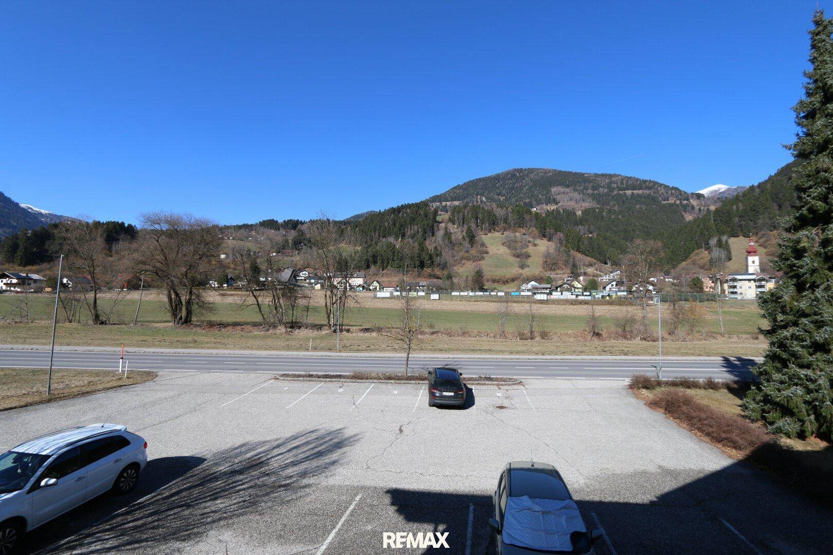 Weitläufiger Blick auf die umliegende Landschaft mit Bergen und einem Parkplatz unter blauem Himmel.