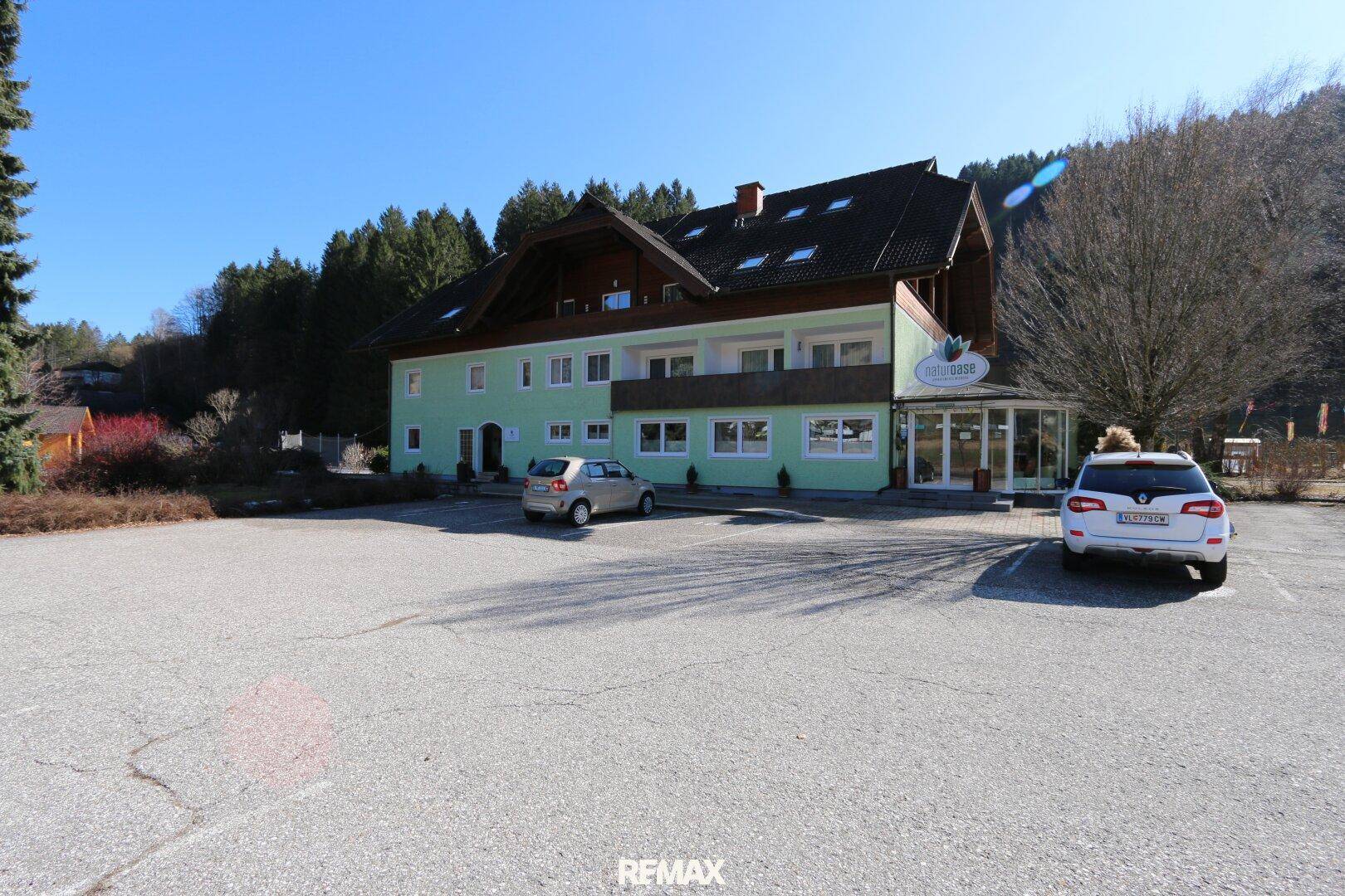 Ansicht eines gepflegten Gebäudes mit Parkplatz und bewaldeter Umgebung unter blauem Himmel.