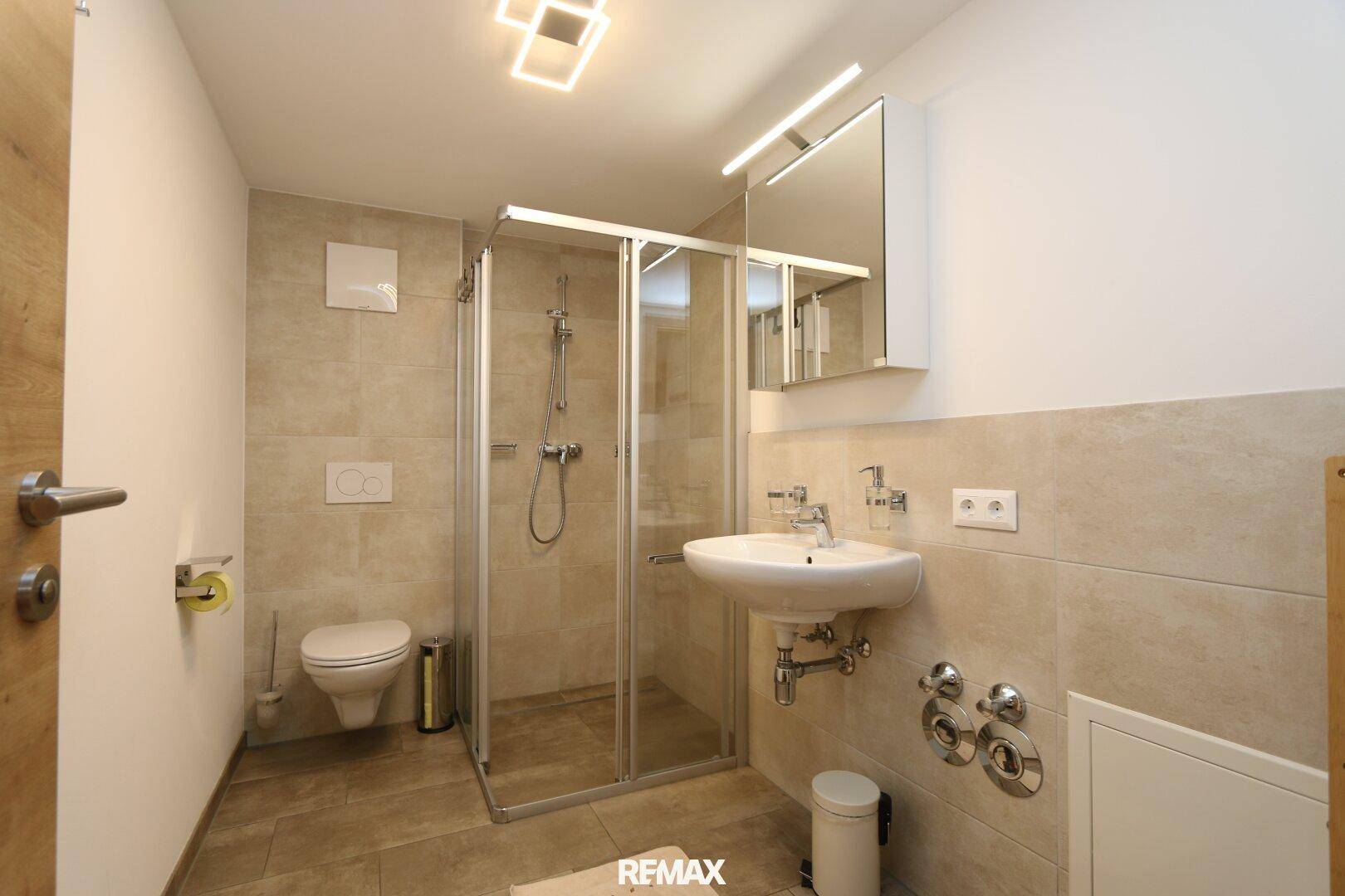 Modernes Badezimmer mit Duschkabine, Wand-WC und Waschbecken in hellen Fliesen.