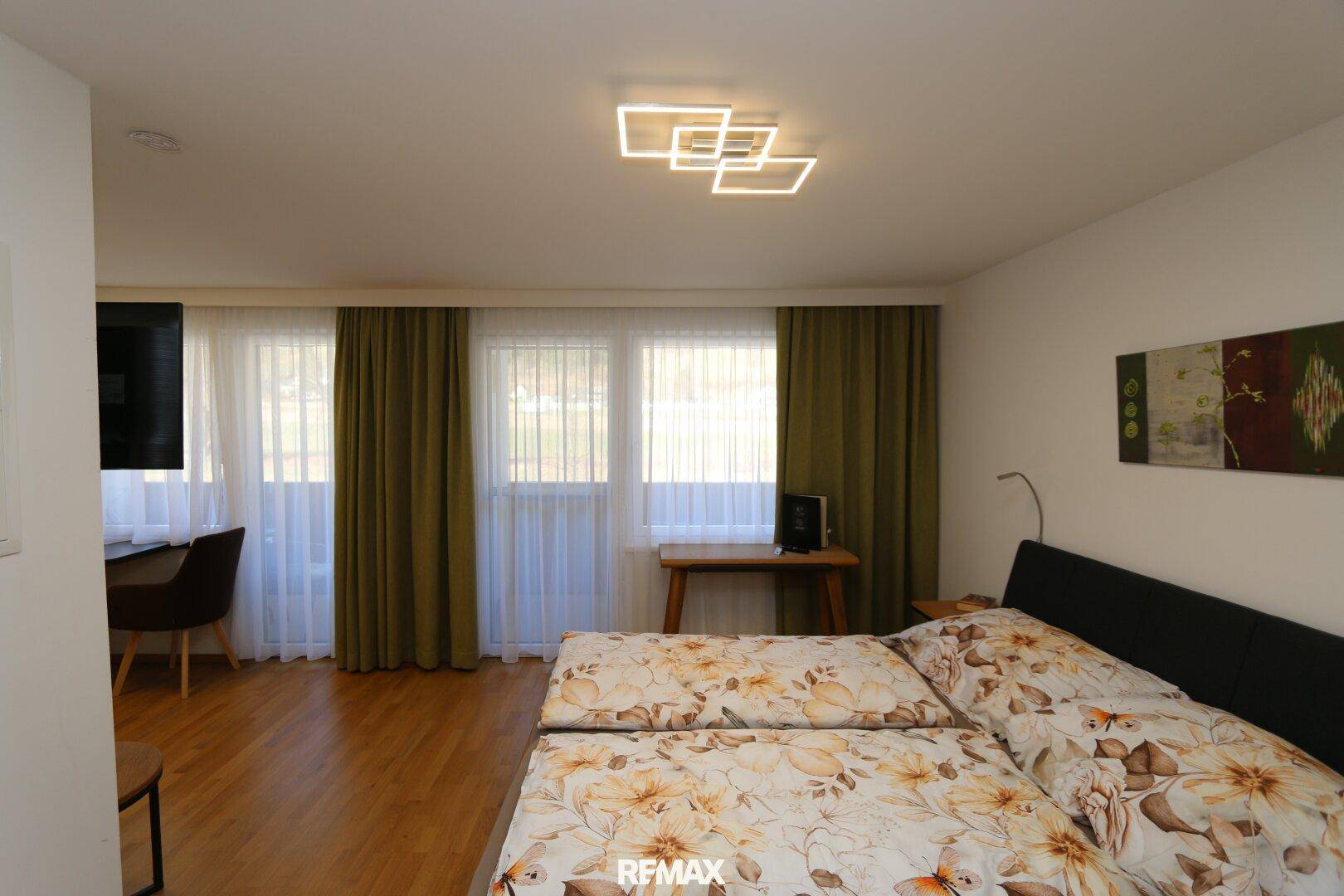 Schlafbereich mit Doppelbett, Holzboden und großen Fenstern, die viel Tageslicht hereinlassen.