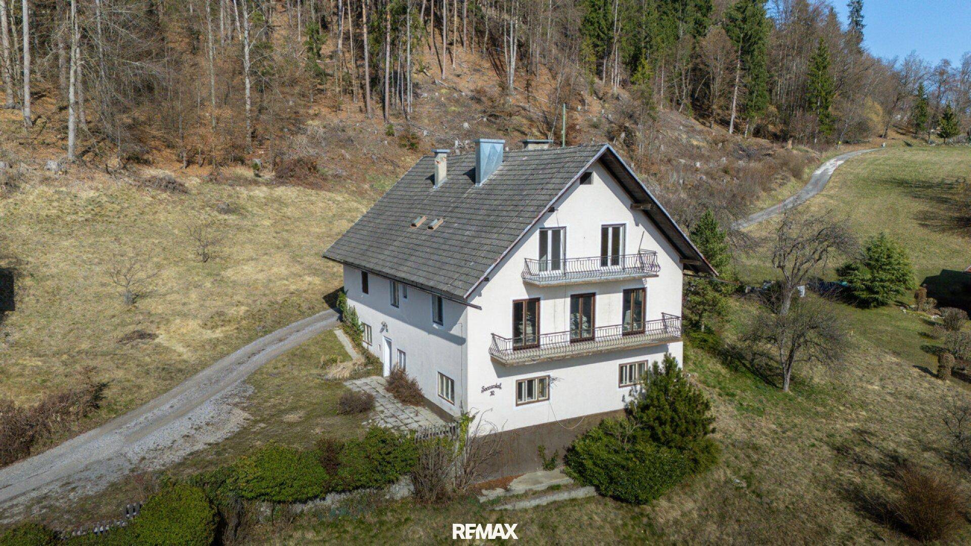 Luftaufnahme des Hauses von der Seite, umgeben von grünen Hügeln und einem Waldgebiet.