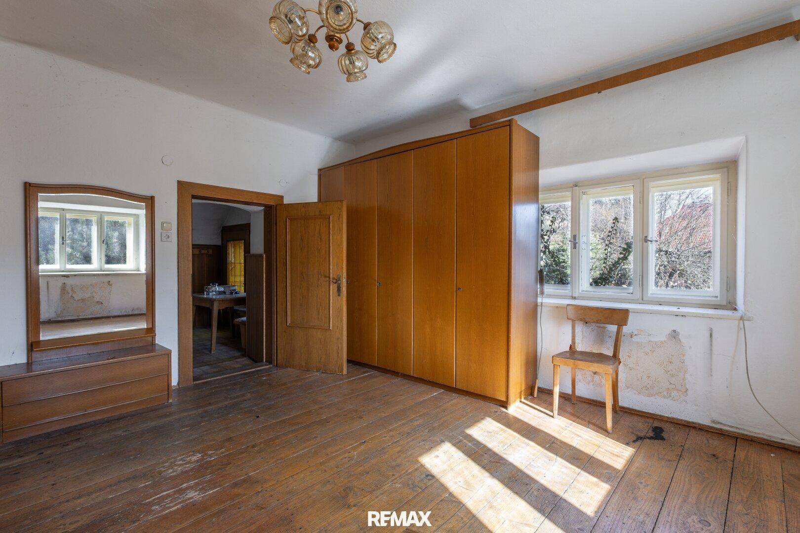 Schlafzimmer mit alten Holzdielen, großem Kleiderschrank und Blick ins Grüne, renovierungsbedürftig.