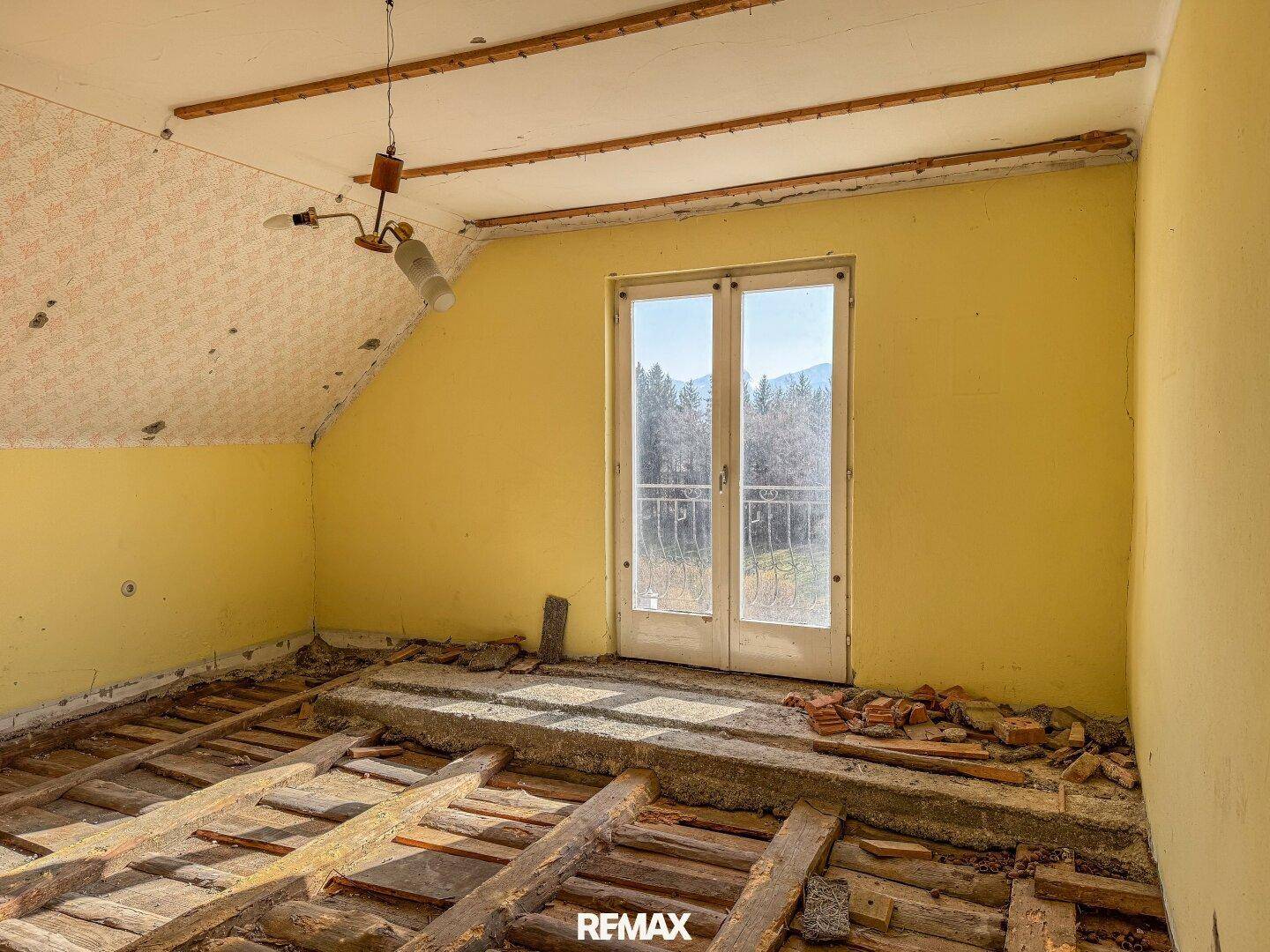 Gelb gestrichenes Dachzimmer im Rohbau mit französischen Balkontüren und weitem Blick auf die Berge.