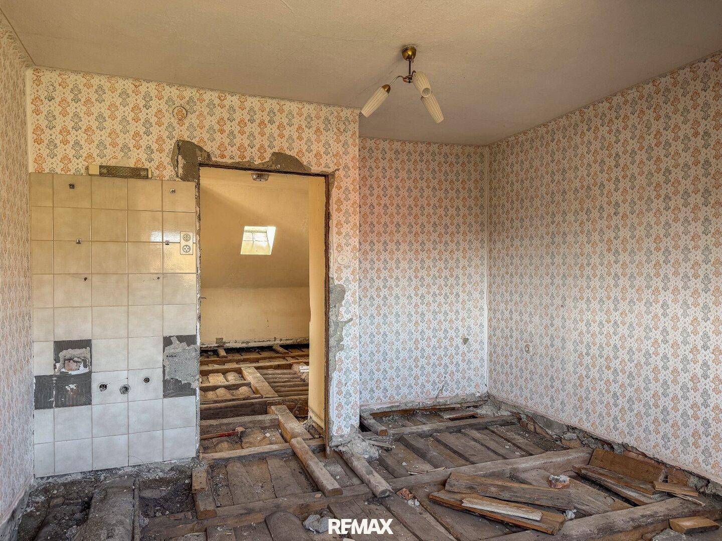 Innenraum im Rohbau mit gemusterter Tapete und freiliegenden Holzbalken, Blick in einen weiteren Raum mit Dachfenster.