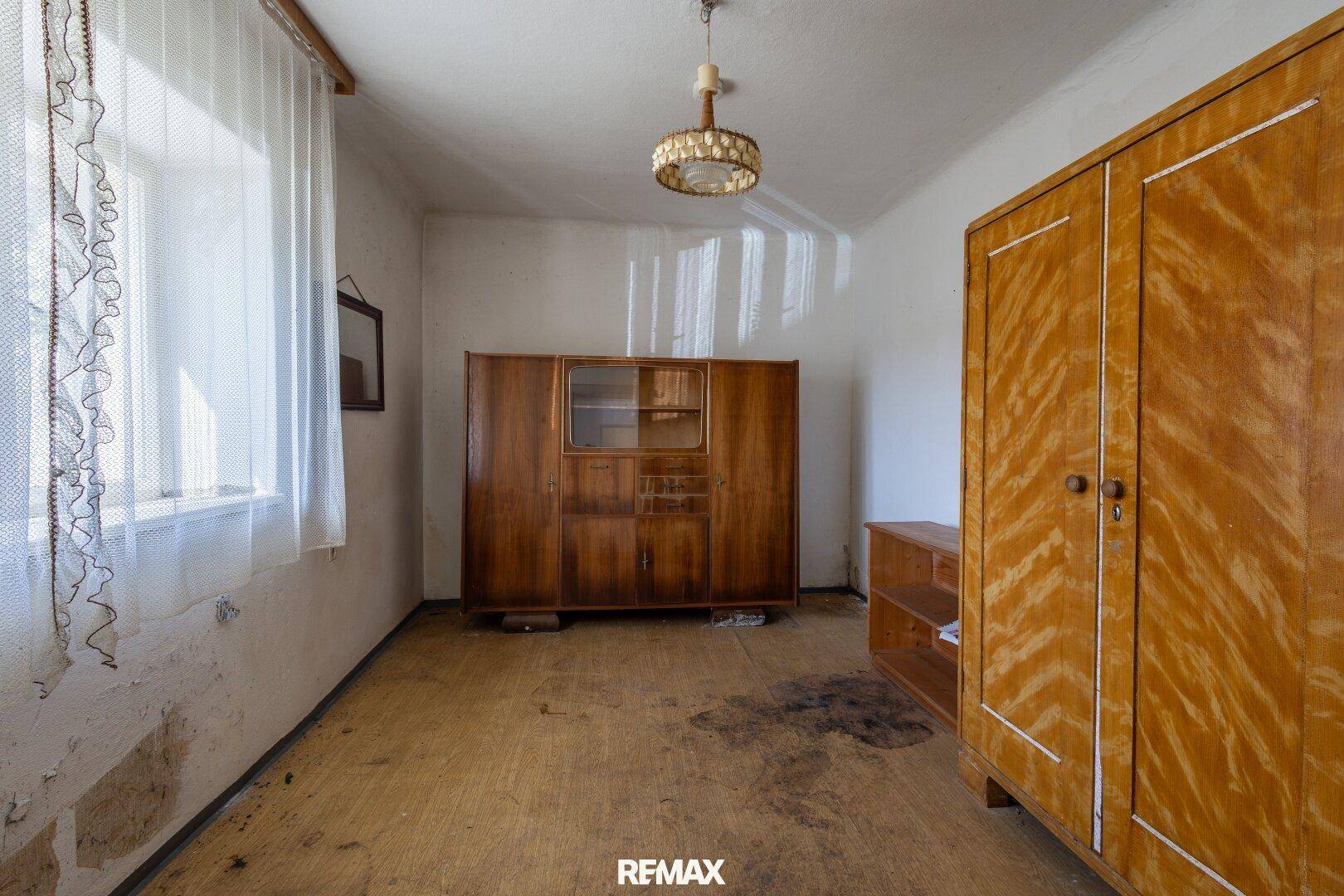 Zimmer mit alten Holzmöbeln und einem Fenster, das viel Tageslicht hereinlässt, in sanierungsbedürftigem Zustand.