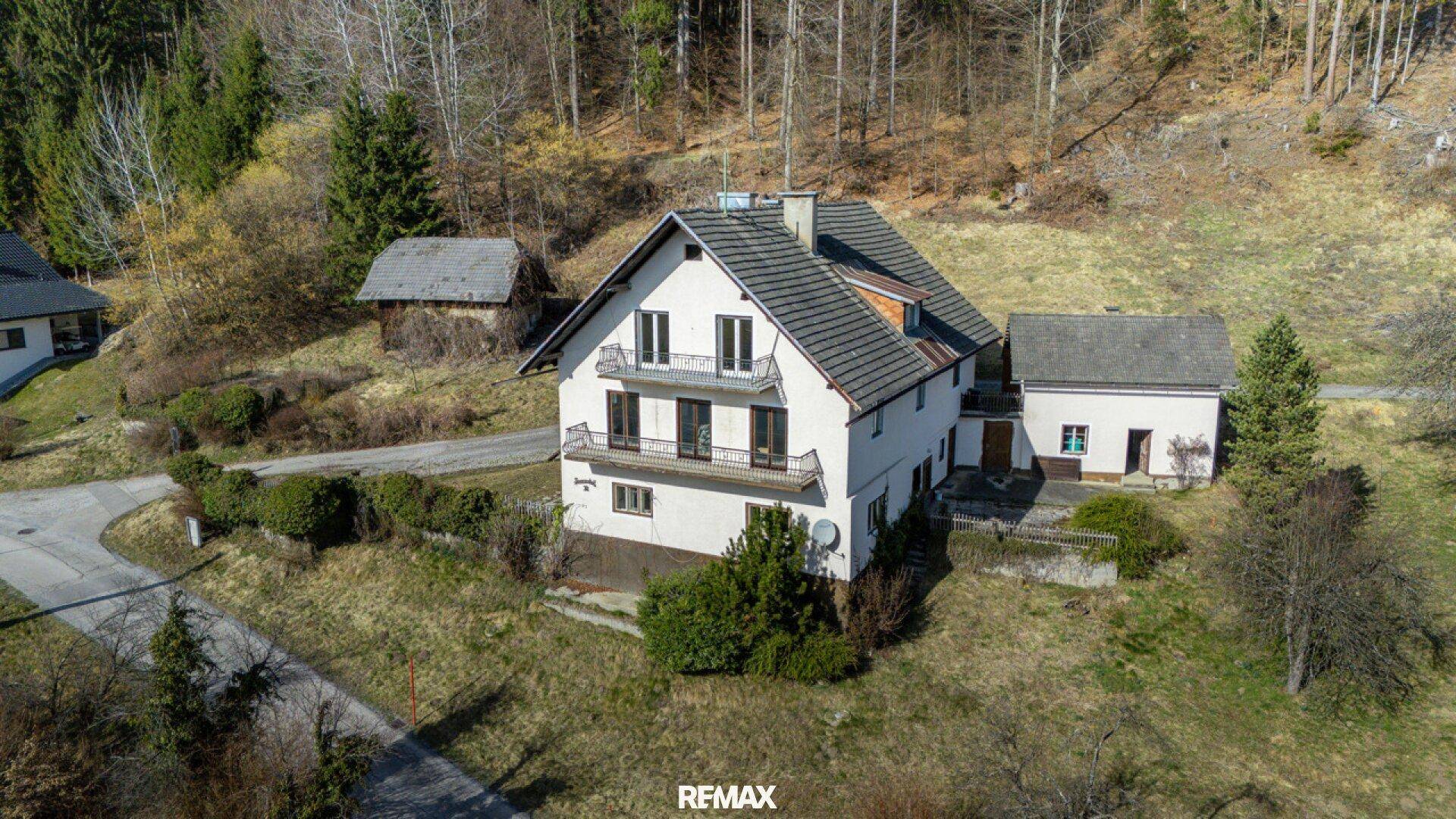Luftaufnahme eines freistehenden Hauses in einer grünen, hügeligen Landschaft mit Wald im Hintergrund.