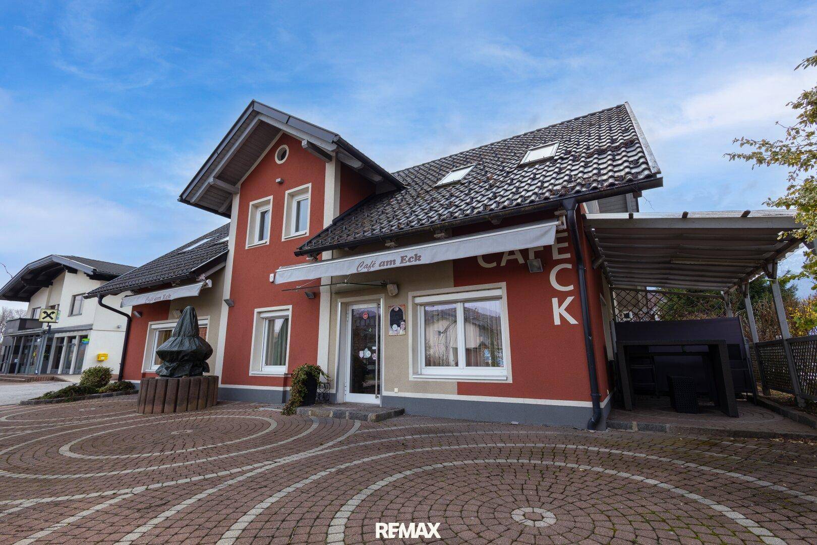 Seitenansicht des Gebäudes mit roter Fassade, Schrägdach und gepflastertem Eingangsbereich.