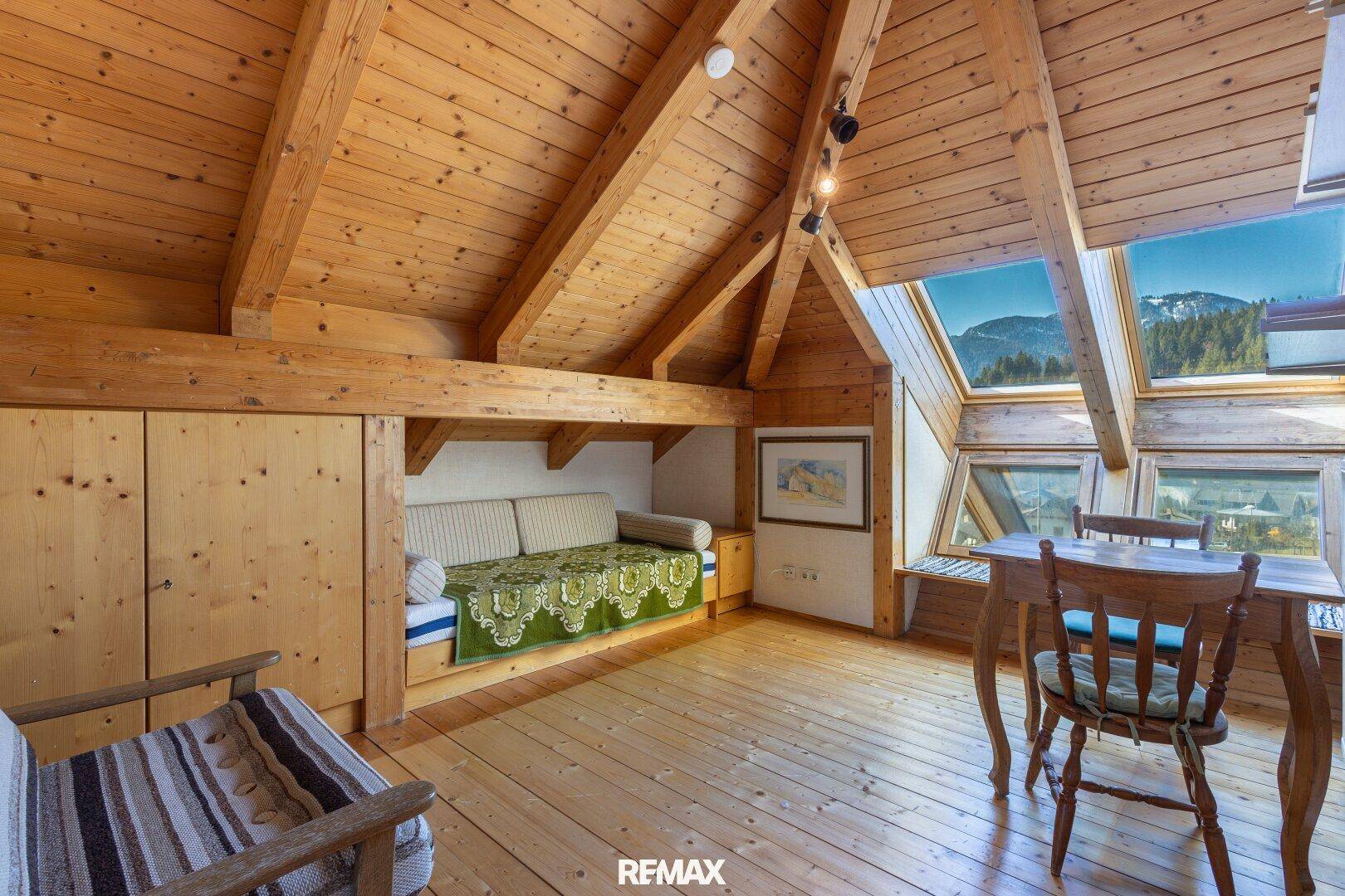Dachzimmer mit Holzbalken, Holzboden und großen Fenstern mit Bergblick.