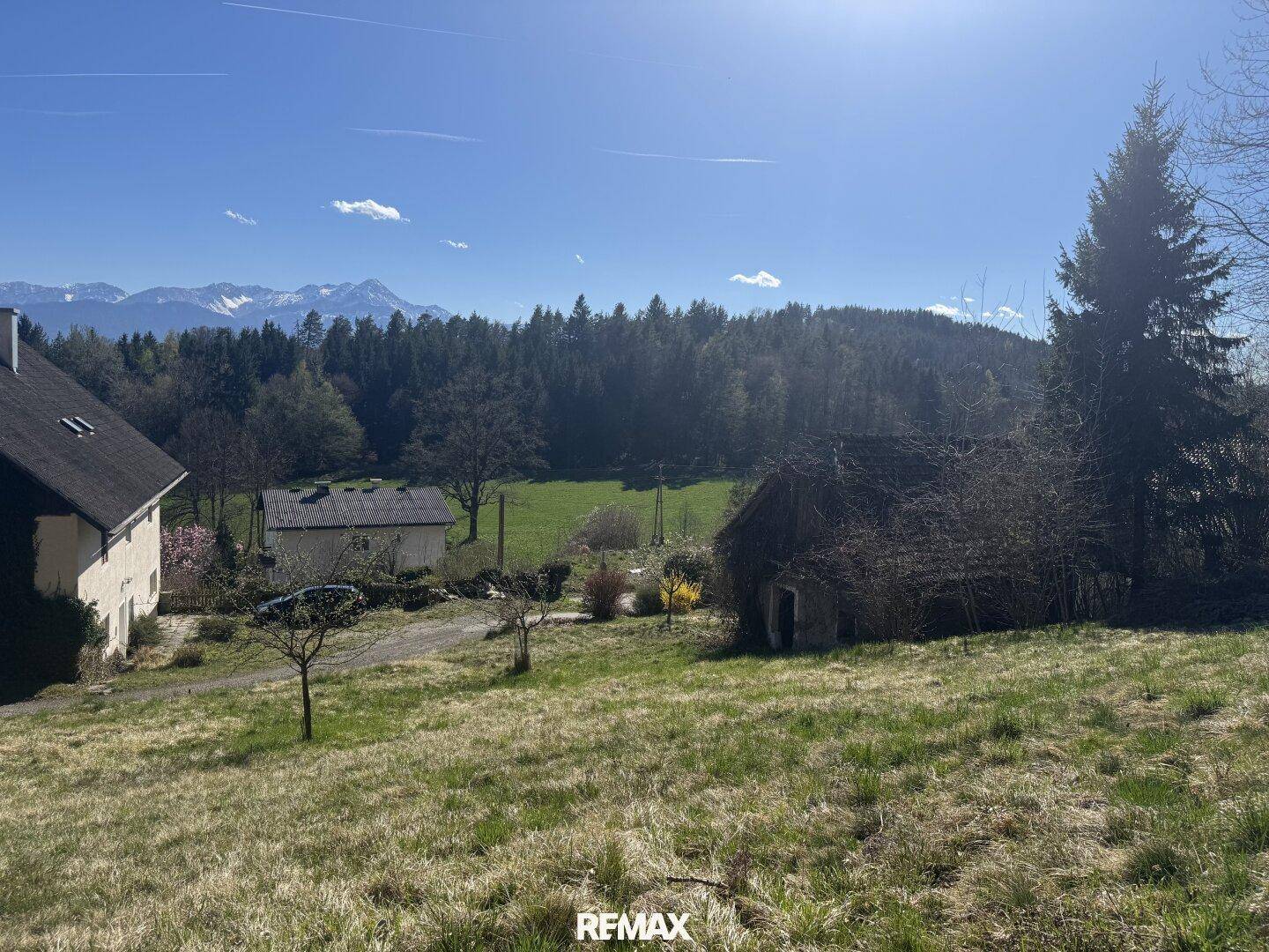 Weitläufige grüne Wiese mit Blick auf bewaldete Hügel und schneebedeckte Berge am Horizont.
