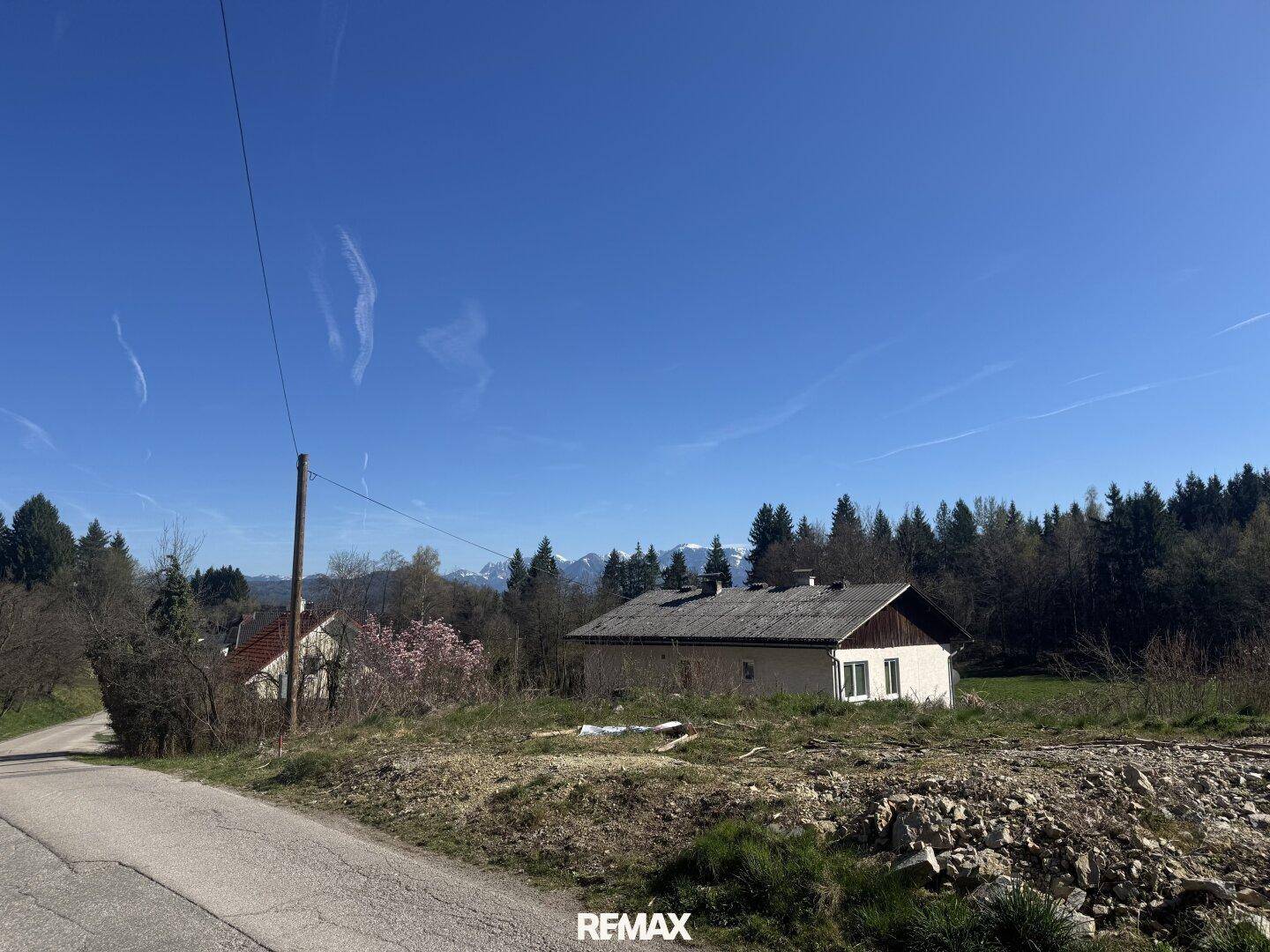 Haus in ländlicher Umgebung an einer Straße, mit Wald und schneebedeckten Bergen im weiten Hintergrund.