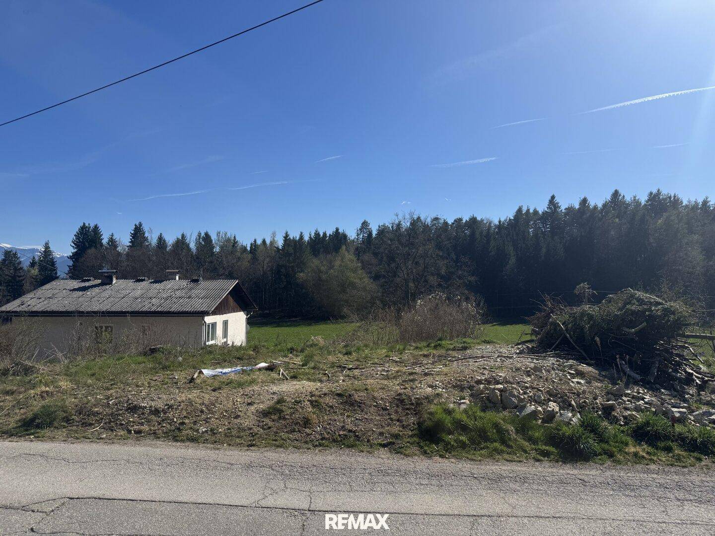 Ländliches Haus mit einfacher Architektur, umgeben von Wiesen und einem Wald, mit Bergen am Horizont.