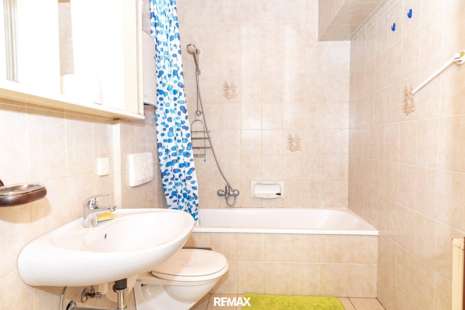 Badezimmer mit beigen Fliesen, Badewanne mit Duschvorhang, WC und Waschbecken.