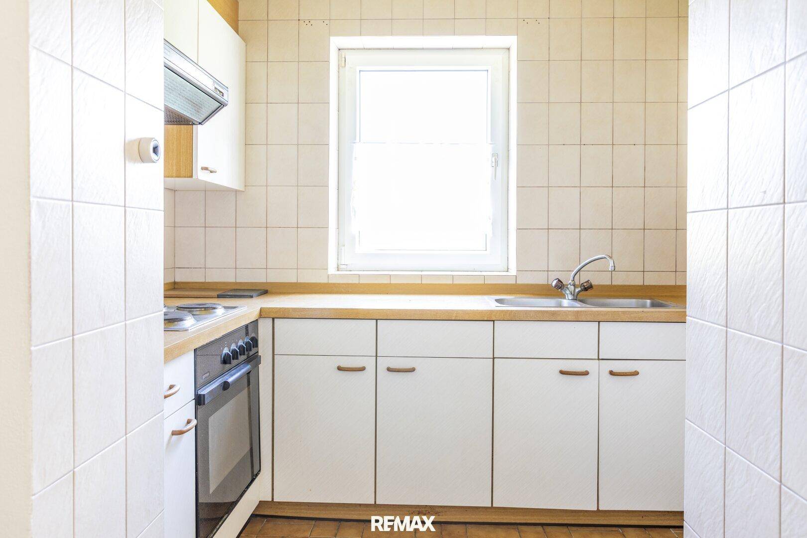 Funktionale Küche mit weißen Schränken, Holz-Arbeitsplatte, Spüle und Fenster für Tageslicht.