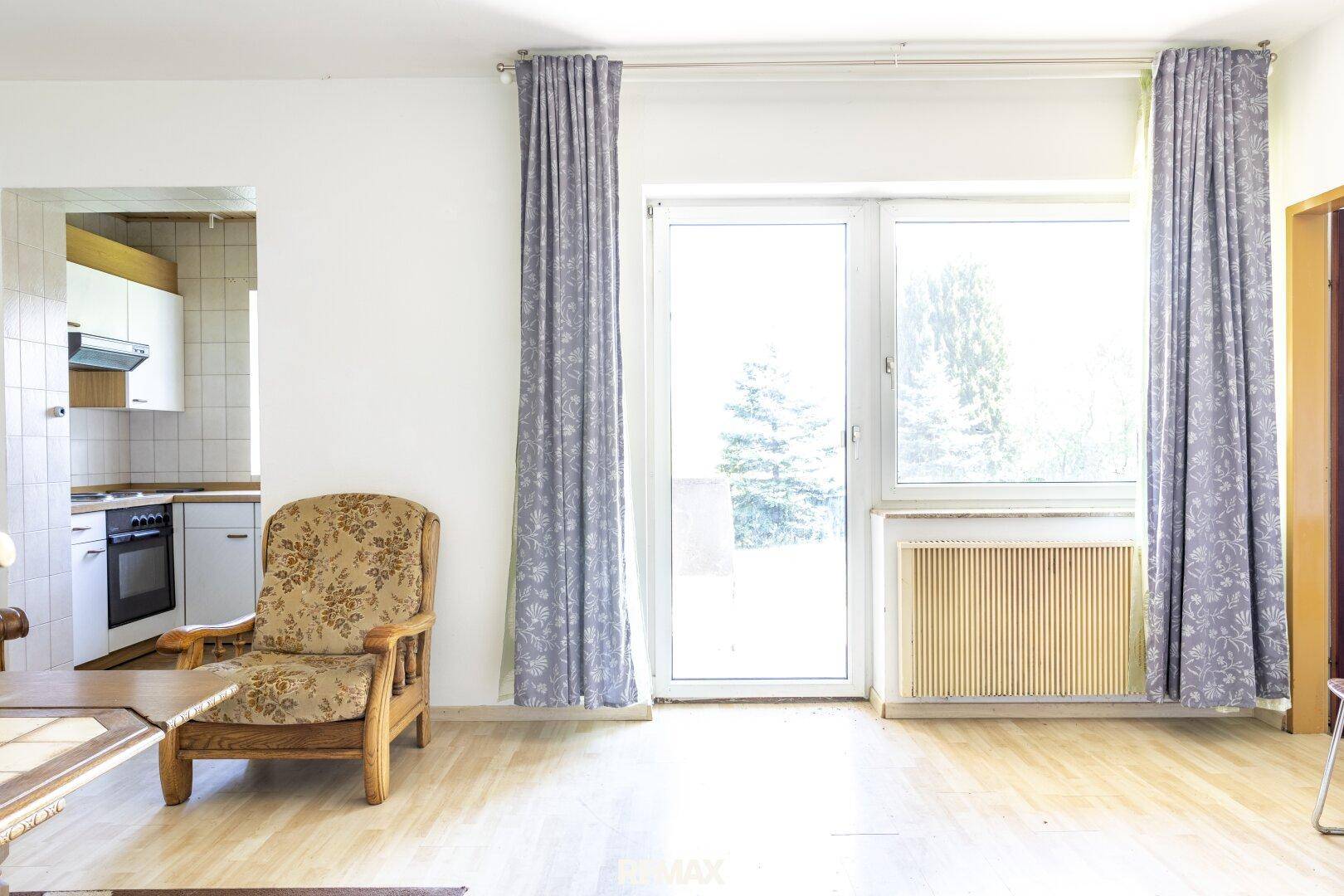 Wohnbereich mit großem Fenster, das viel Licht hereinlässt, und Blick in die angrenzende Küche.