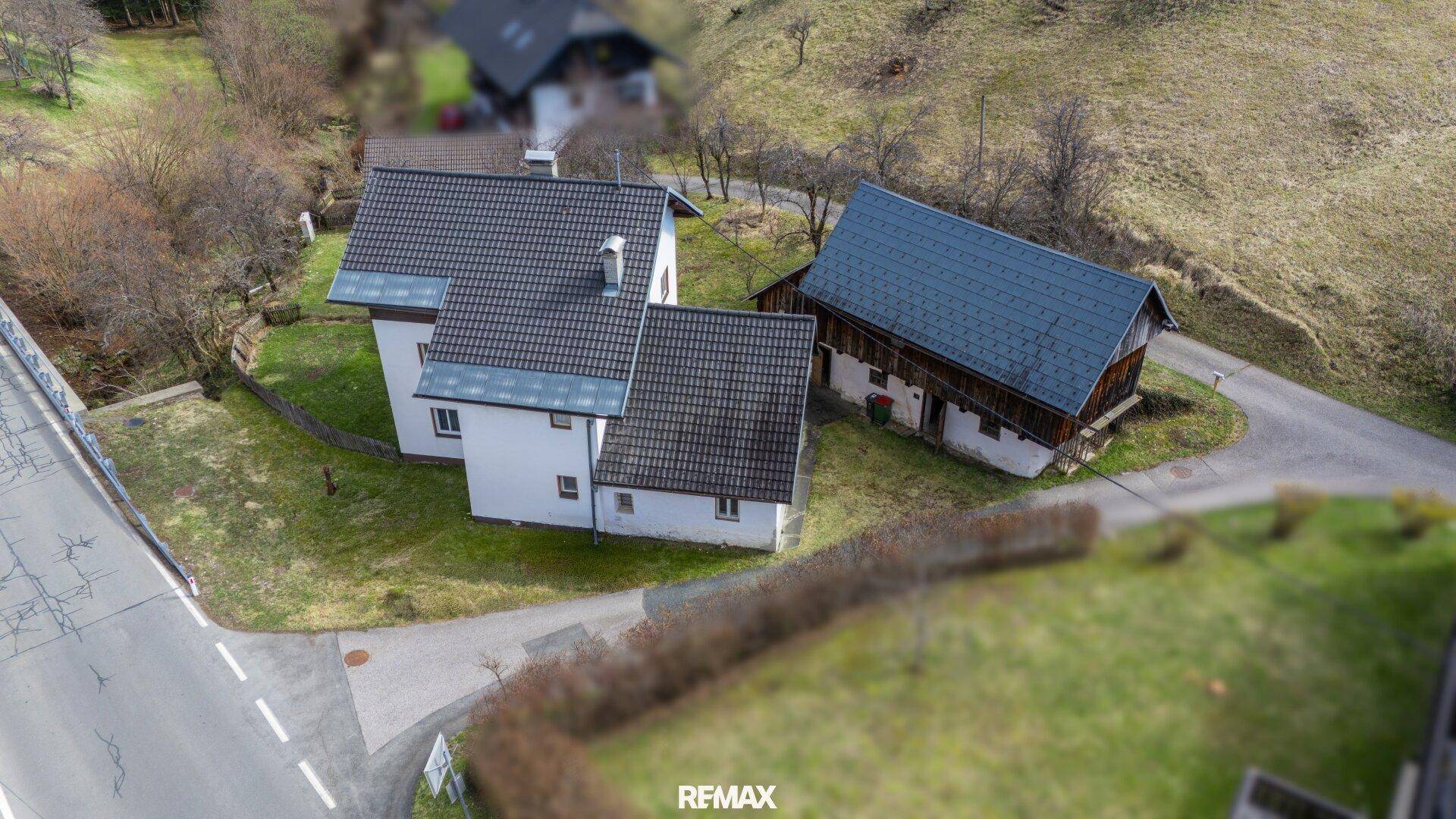 Luftaufnahme des Anwesens mit Haupthaus und angrenzender Scheune in hügeliger Landschaft.
