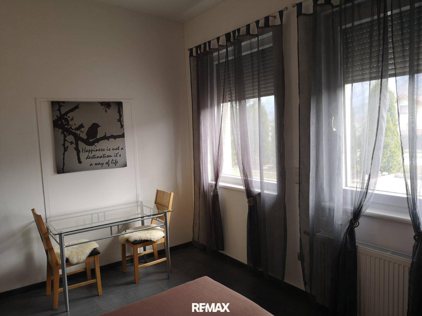 Gemütliches Schlafzimmer mit kleinem Tisch, Stühlen und zwei Fenstern mit dunklen Vorhängen.